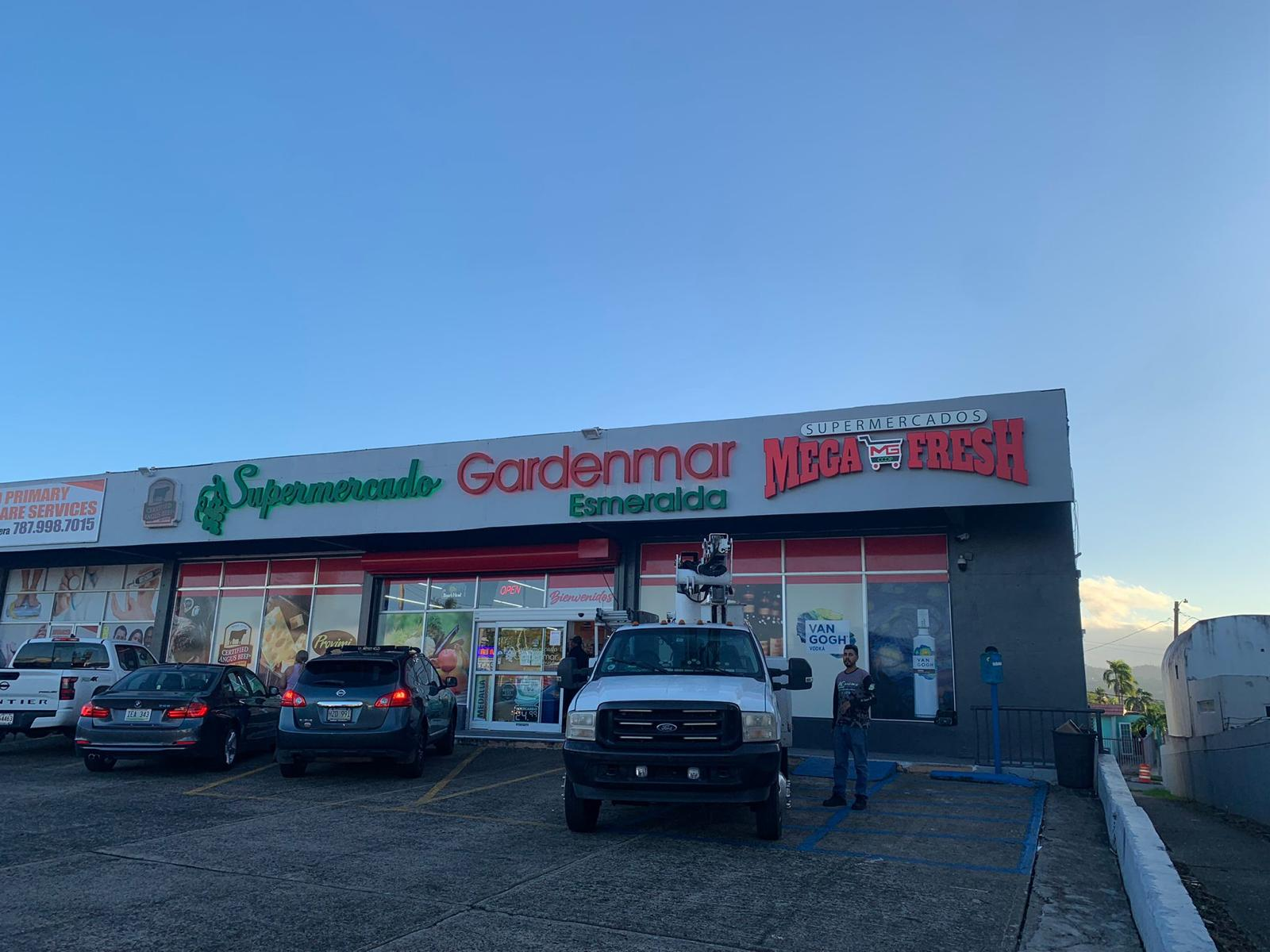 Fachada de un supermercado llamado 'Supermercado Gardenmar Esmeralda', con varias marcas y logotipos, incluyendo 'Mega Fresh'. Hay autos en el estacionamiento y dos personas frente a la tienda.