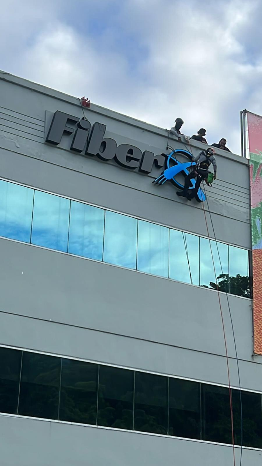 Personal instalando la señal de un supermercado llamado 'Fifer' en un edificio moderno con ventanas de vidrio, mientras algunas personas observan desde la azotea, en un día nublado.