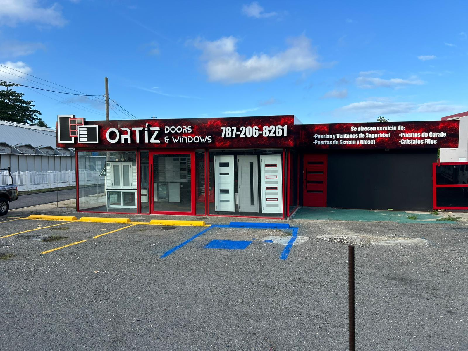 fachada de tienda de puertas y ventanas, con letrero en rojo y negro que dice "ORTiz DOORS & WINDOWS" y un número de teléfono, con exhibición de puertas, ventanas y cristales en el interior, estacionamiento en primer plano.