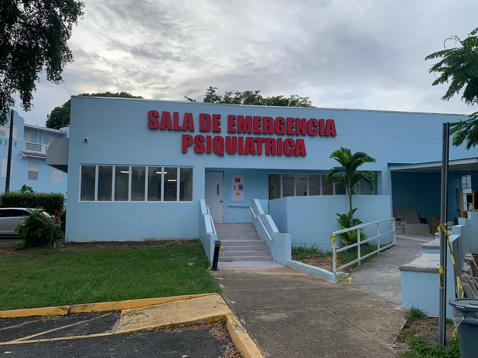 Edificio con cartel que dice 'Sala de Emergencia Psiquiátrica', en color rojo, en una clínica o hospital, rodeado de áreas verdes y con escaleras de acceso