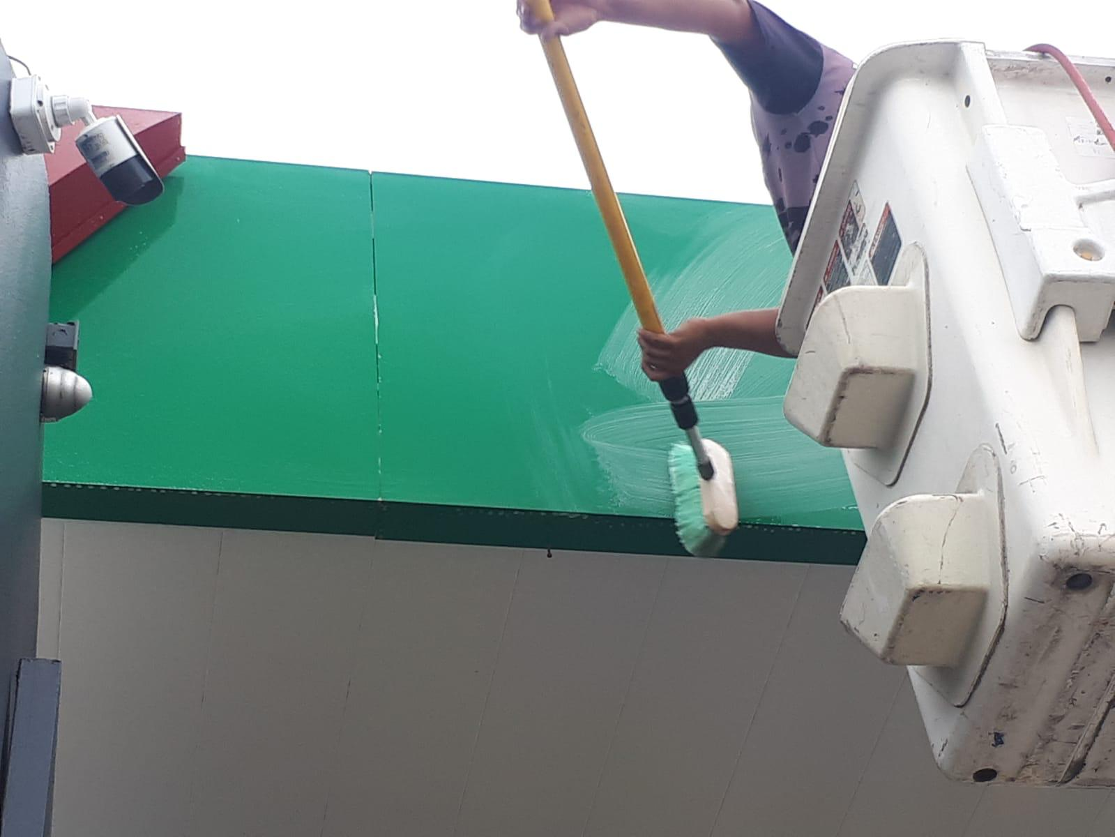 Una persona en una plataforma de trabajo lavando un cartel grande de color verde con una manguera y cepillo de limpieza.
