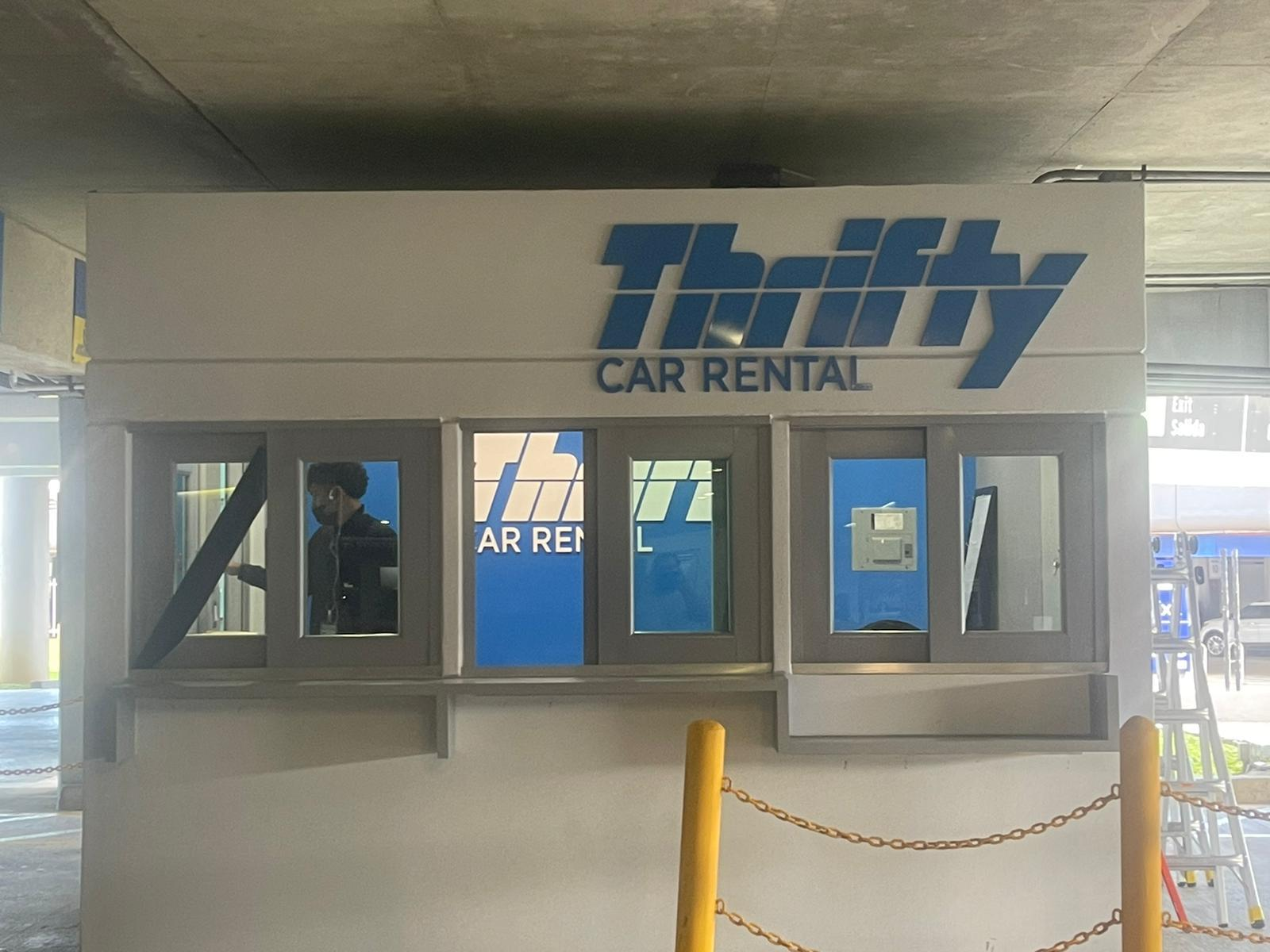 Mostrador de alquiler de autos con señal que dice 'Thrifty Car Rental', ventanas transparentes, una persona usando audífonos y una escalera a la derecha.