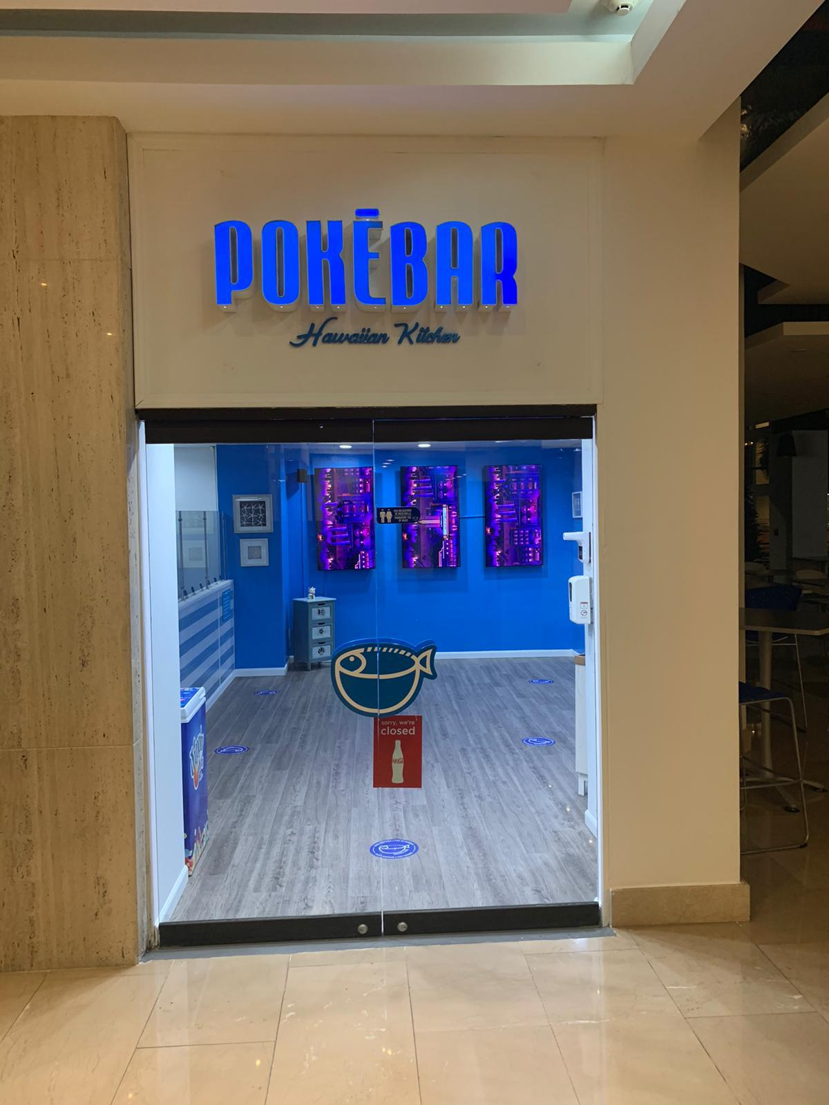 Entrada de un establecimiento llamado 'Pokébar' con decoración en tonos azules, exhibiendo un cartel con un pez y señal de cerrado en la puerta. El interior tiene paredes azules, arte en la pared y un ambiente moderno.