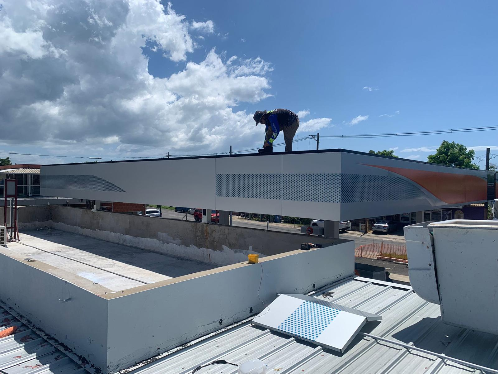 Un trabajador en casco y ropa de protección colocando lo que parece ser paneles decorativos en la fachada de un edificio en construcción, con cielo parcialmente nublado de fondo.