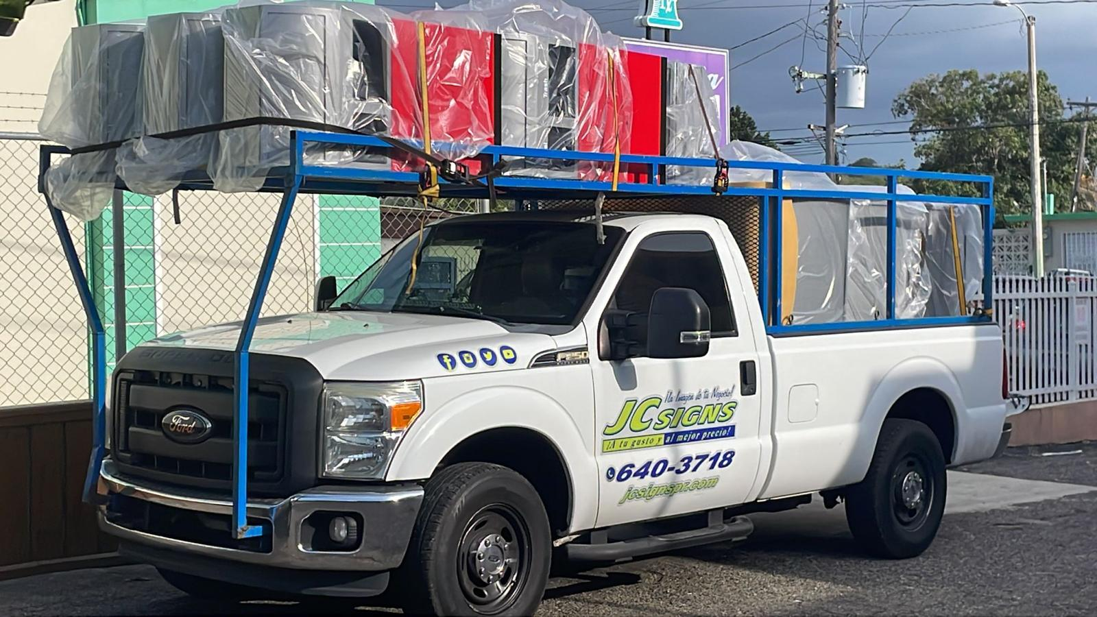 Camioneta blanca con publicidad de JC Signs en la puerta y estructura metálica azul en la parte superior, transportando paquetes envueltos en plástico, en una calle con fondo de cielo nublado y cables eléctricos.