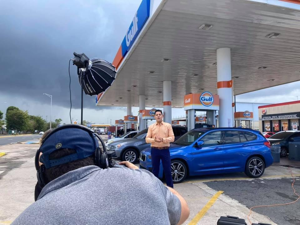 Una escena en una gasolinera Gulf donde un hombre está posando frente a una cámara, mientras un camarógrafo ajusta su equipo de grabación, con un coche azul estacionado al fondo y un cielo nublado.