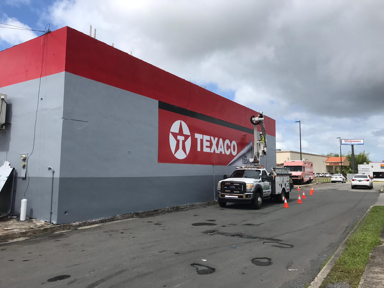 Pintura en la pared de un edificio de la marca Texaco, con un camión que pinta la pared y conos de seguridad a su alrededor. Al fondo, signos de tiendas Walgreens y otros comercios.