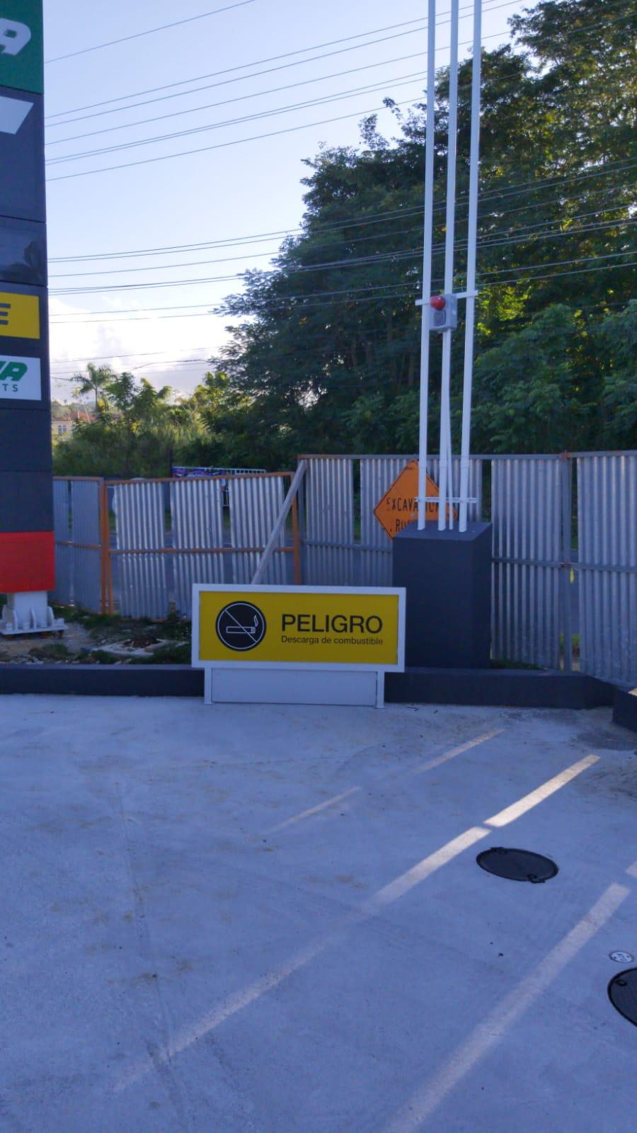 Señal de peligro por descarga de combustible en un estacionamiento, con una barrera simula un tren y valla de seguridad en el fondo.