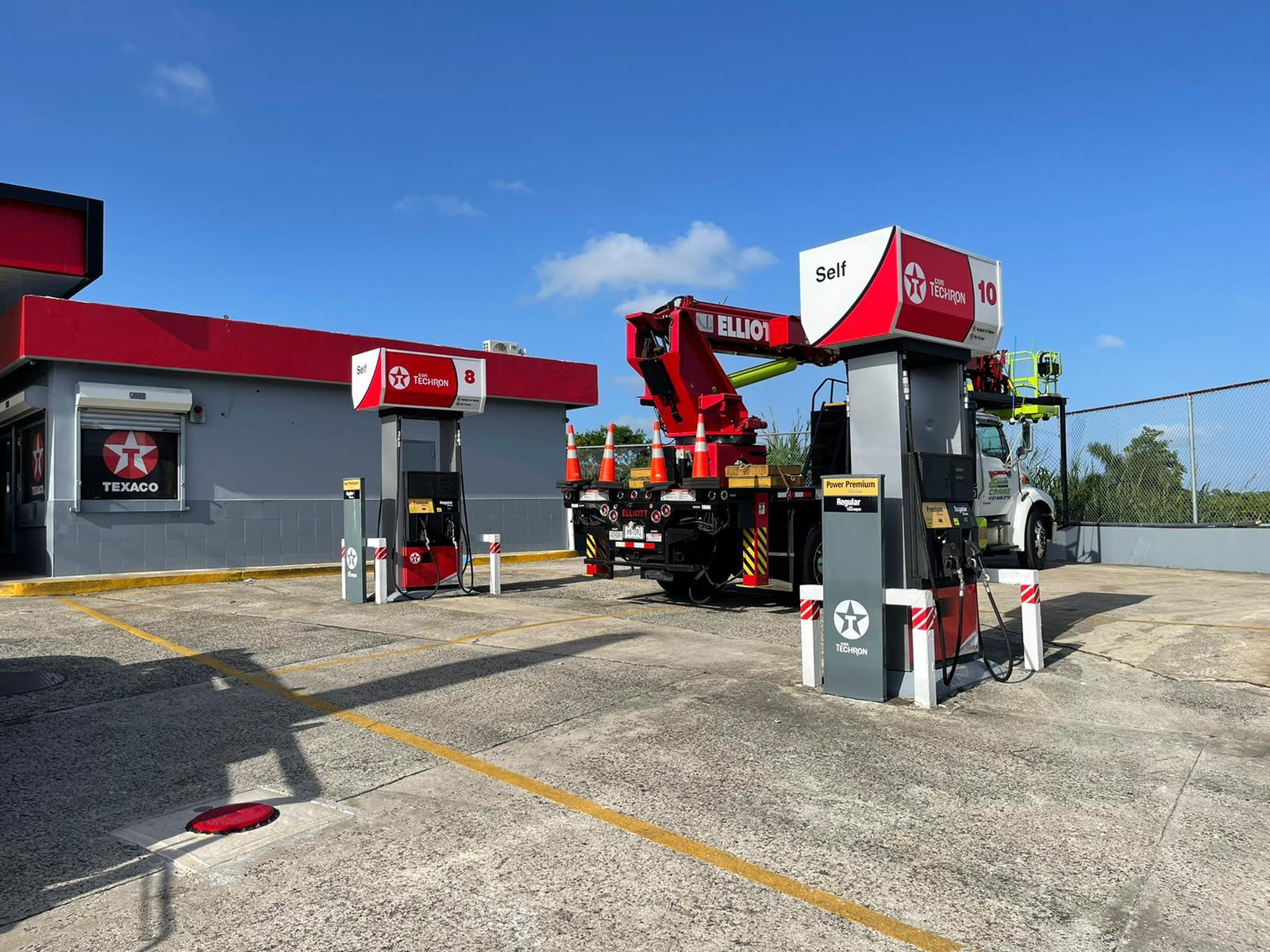 Estación de servicio con dos bombas de gasolina de marca Texaco, una de ellas con servicio de autoservicio, y un camión con equipo de mantenimiento en el fondo, bajo un cielo despejado.