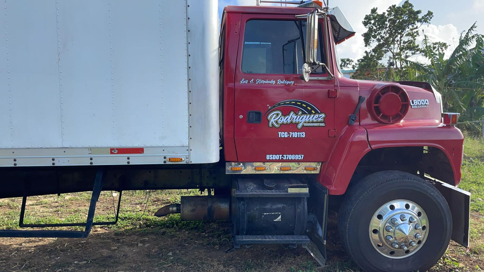 Camión de transporte de carga de color rojo y blanco, con el logotipo de Rodriguez y número de identificación, estacionado en un campo con árboles y césped.