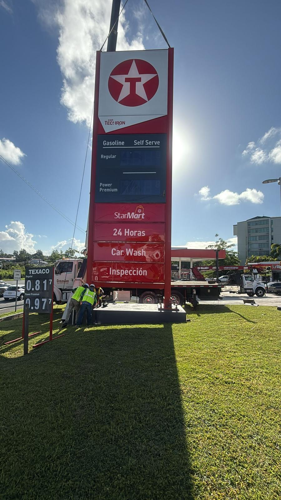 Una torre de señalización de una estación de servicio con información sobre gasolina, autoservicio, 24 horas, lavado de autos e inspección, y un grupo de personas trabajando en la base.