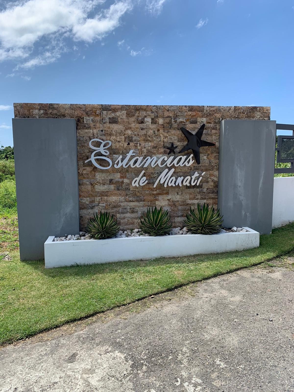 Cartel de bienvenida que dice 'Estancias de Manatí' con estrellas y plantas en un macetero.