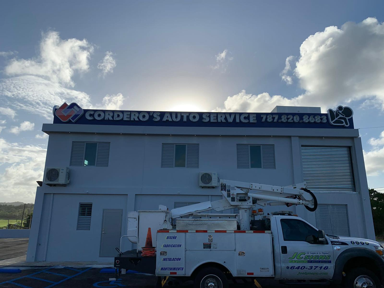 Fachada de un taller de autos con cartel que dice 'Cordero's Auto Service' y teléfono, con un camión de trabajo en primer plano.