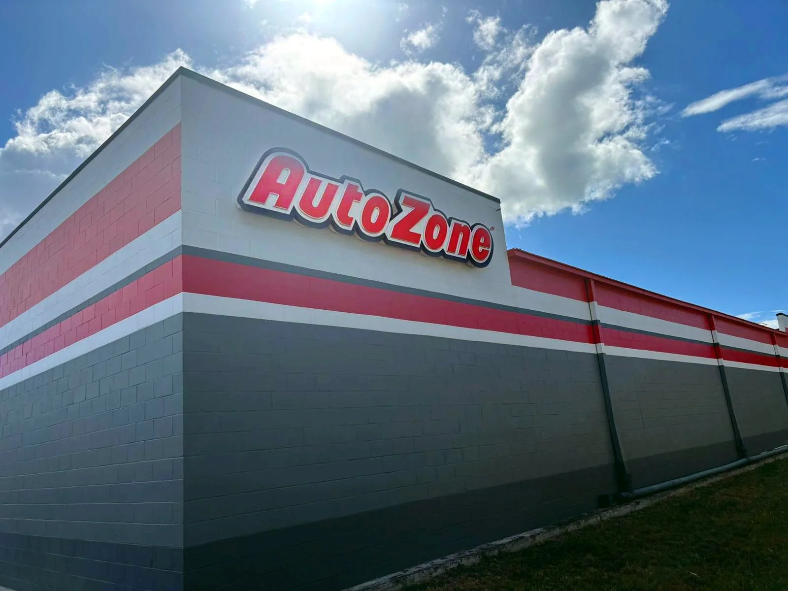 Edificio de tienda con cartel que dice AutoZone, con paredes pintadas en colores gris, rosa y blanco, bajo un cielo con nubes y sol.