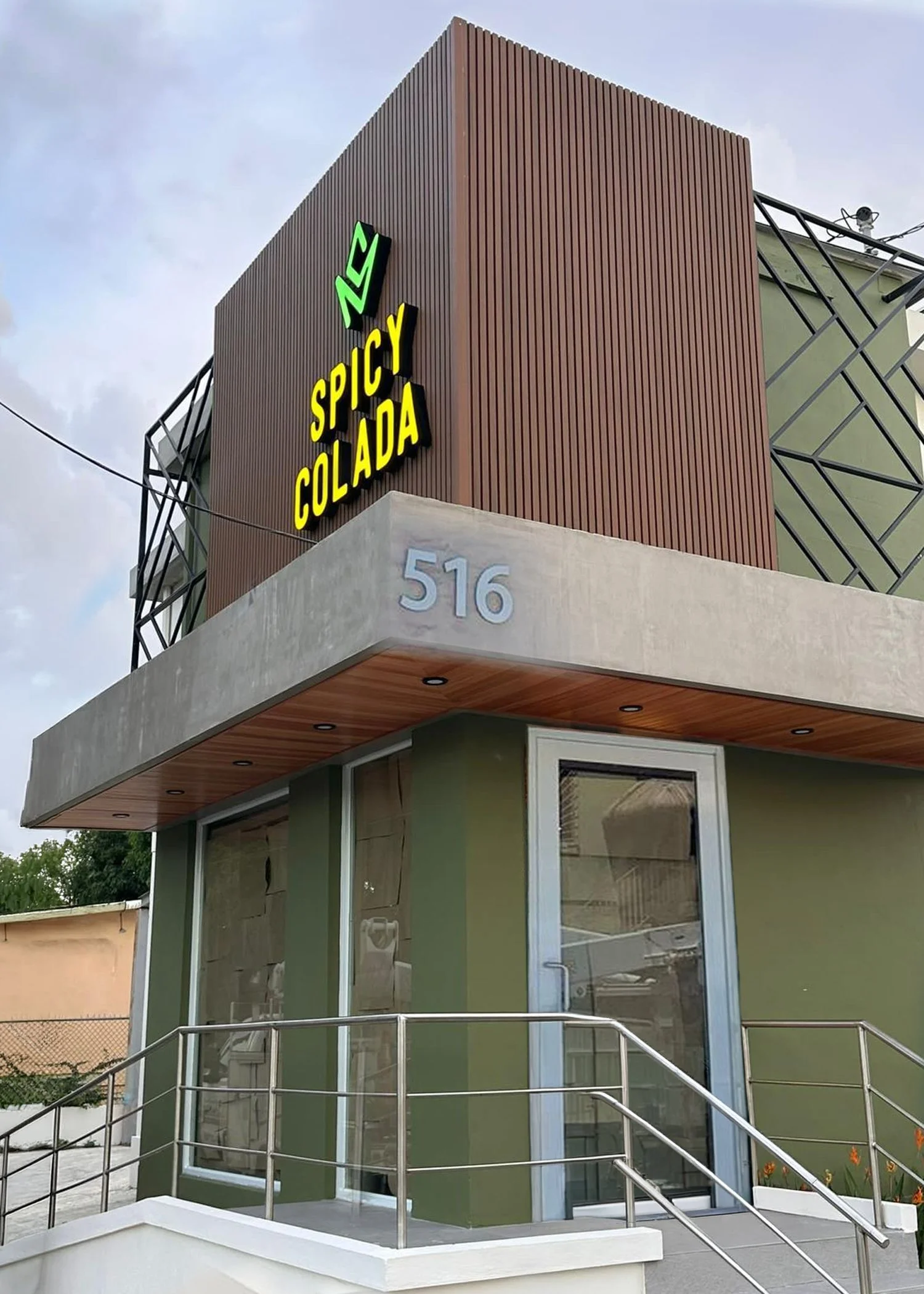 Fachada de un edificio moderno con letrero que dice 'Spicy Collada' en letras de colores neón y número 516 en la pared, con un entorno con plantas y estructura de barandales de acero inoxidable.