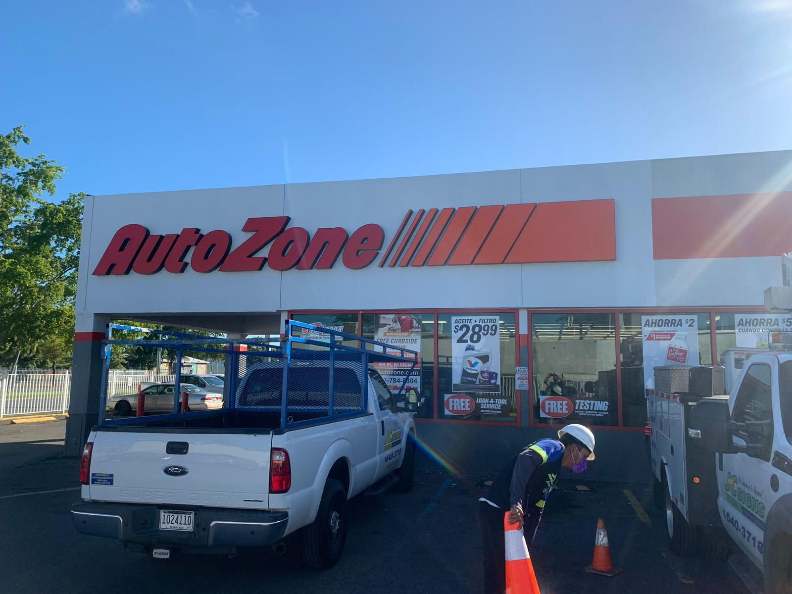 Fachada de un edificio de Auto Zone con vehículos y empleados en el estacionamiento, algunos realizando tareas de trabajo con conos y equipo de protección.