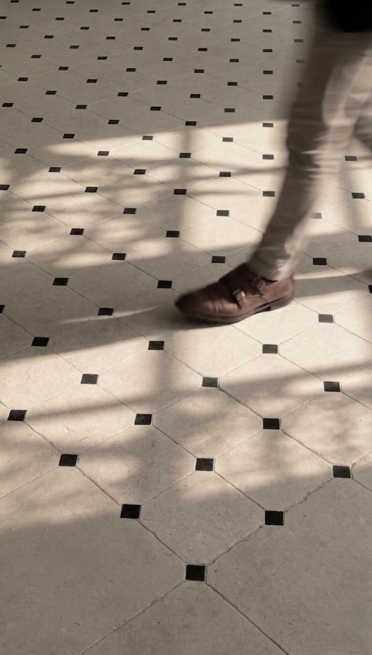 A person's legs and brown shoes walking on a tiled floor with a pattern of black squares and octagonal tiles, with shadow patterns cast across the floor.