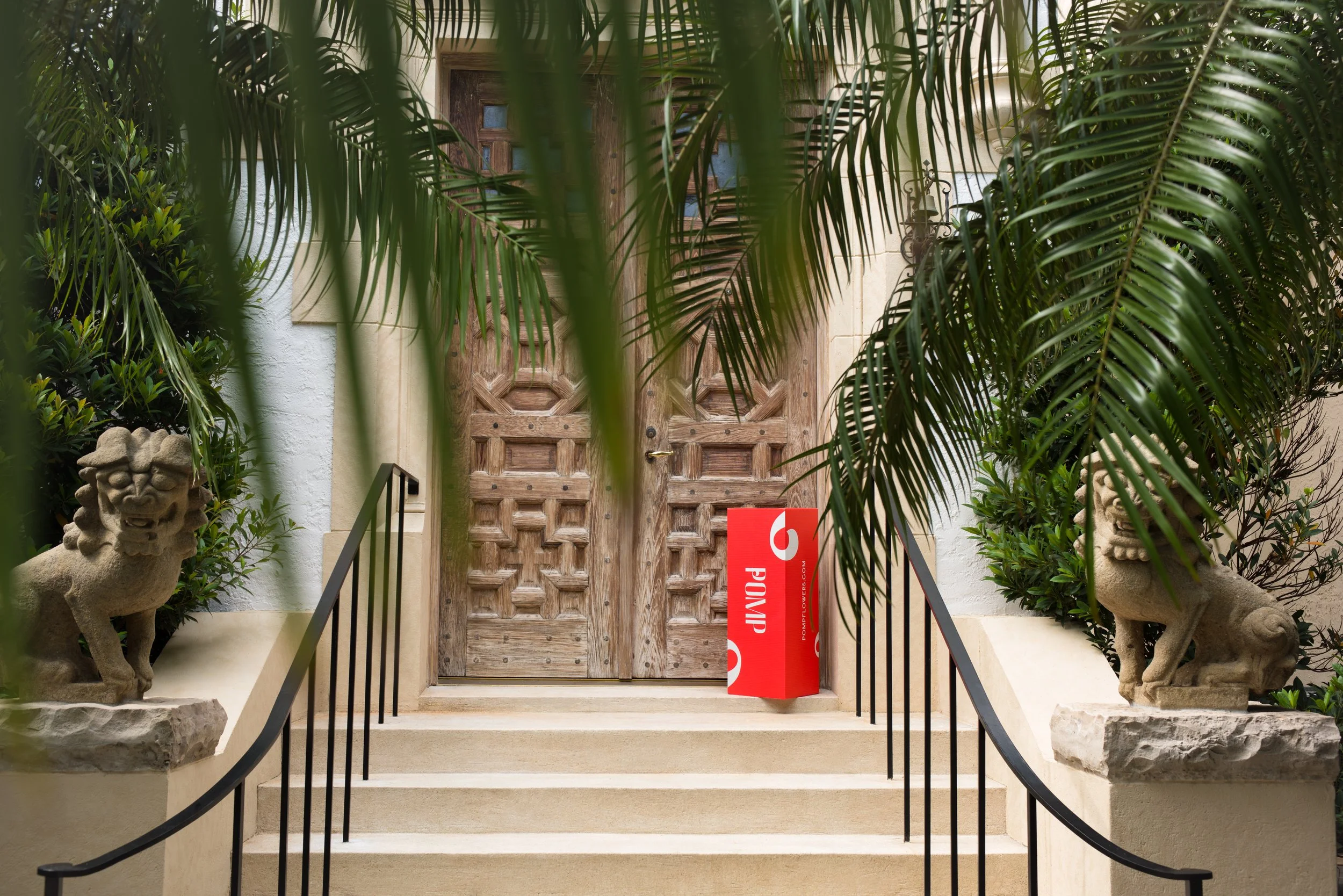 Wooden front door with small windows, flanked by garden statues of lions, surrounded by lush green tropical plants, stairs with black railings, and a red 'Open' sign.