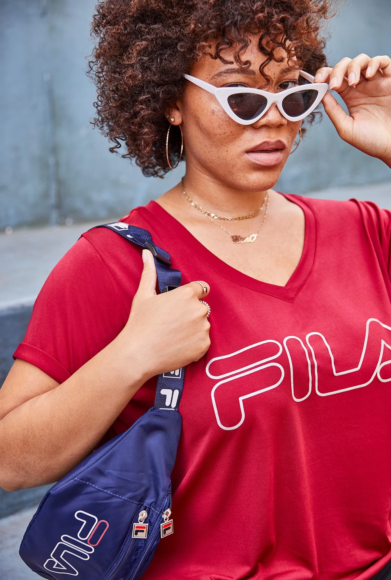 A woman with curly hair wearing white cat-eye sunglasses, a red FILA t-shirt, gold jewelry, and carrying a navy FILA backpack.