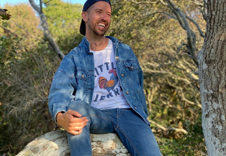 A man with a beard and mustache sitting on a large rock outdoors, smiling. He's wearing a denim jacket, a white T-shirt with a rooster graphic and text, and a backward baseball cap. Trees with green and yellow leaves are in the background.