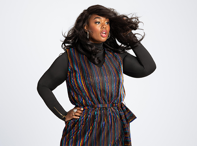 A woman with long, curly dark hair wearing a colorful, striped dress with a black turtleneck underneath, posing against a plain white background.