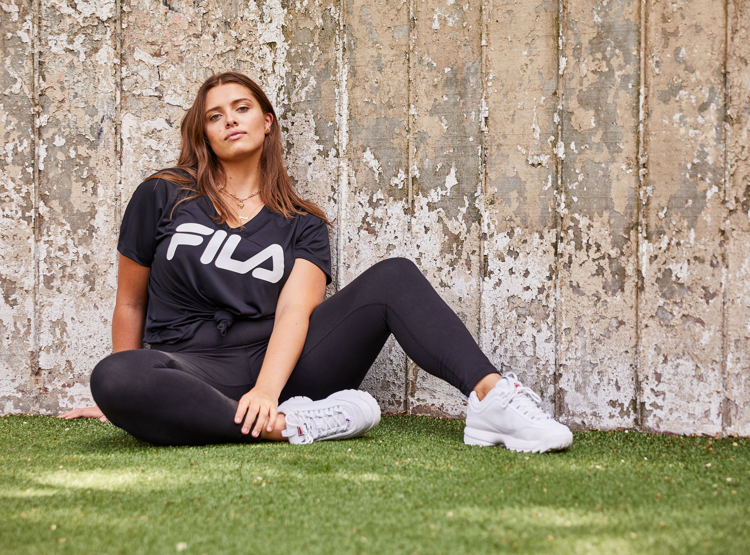 A young woman with brown hair sitting on the grass against a weathered wall with peeling paint, wearing a black FILA T-shirt, black leggings, and white sneakers, looking confidently at the camera.