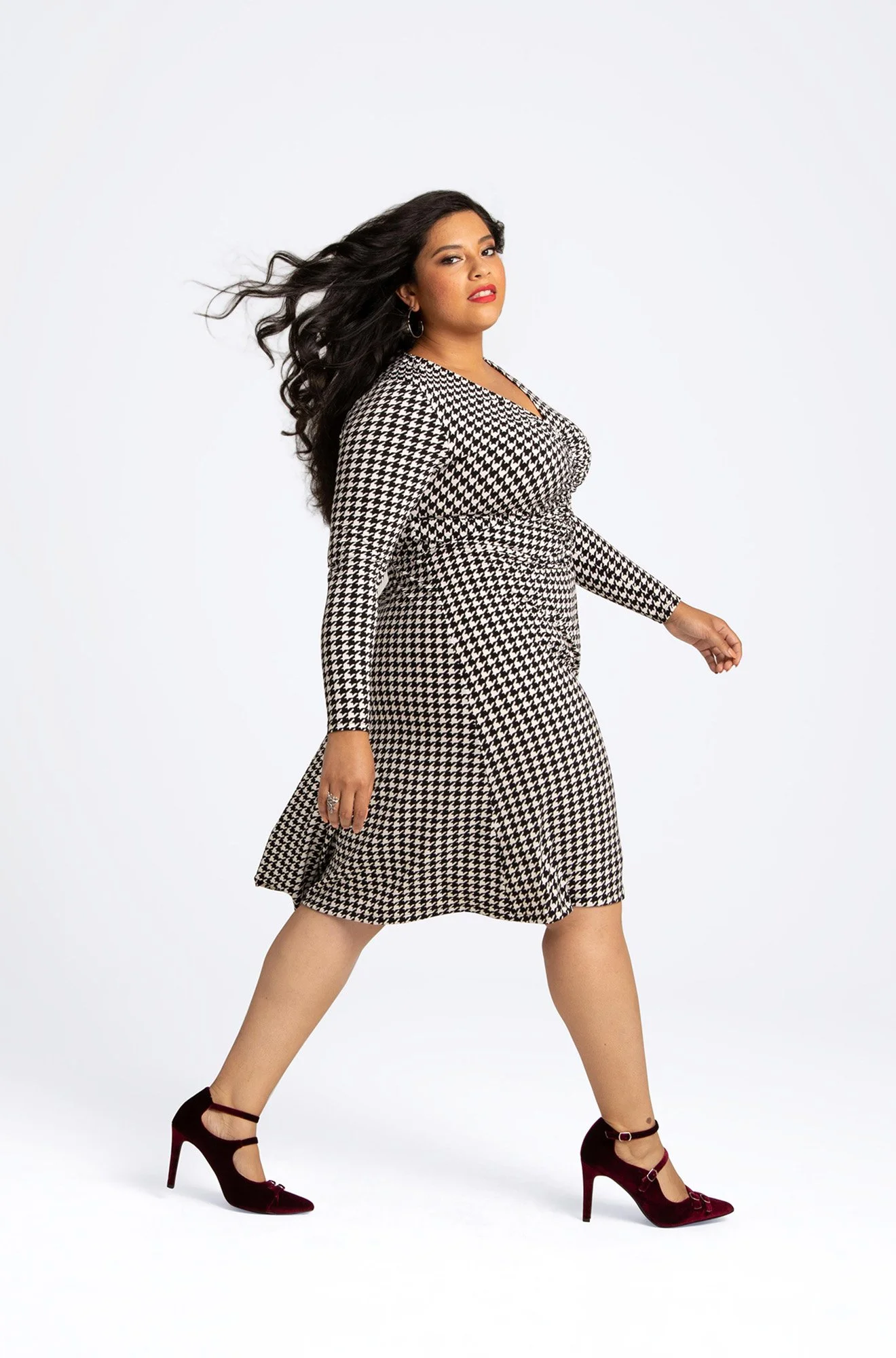 A woman in a black and white houndstooth patterned dress and maroon high heels is walking confidently, with her long dark hair flowing and a slight smile on her face.