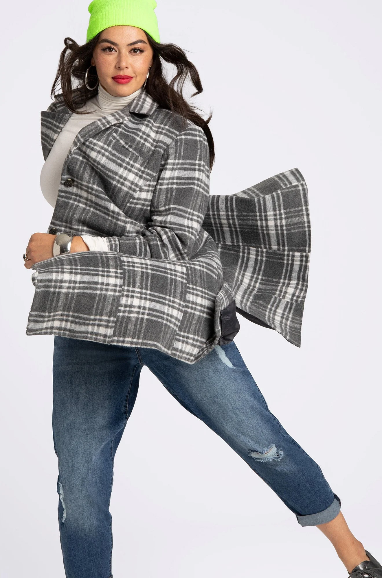 A woman wearing a neon green beanie, a white turtleneck, a gray plaid coat, and ripped jeans poses with her legs apart against a plain background.