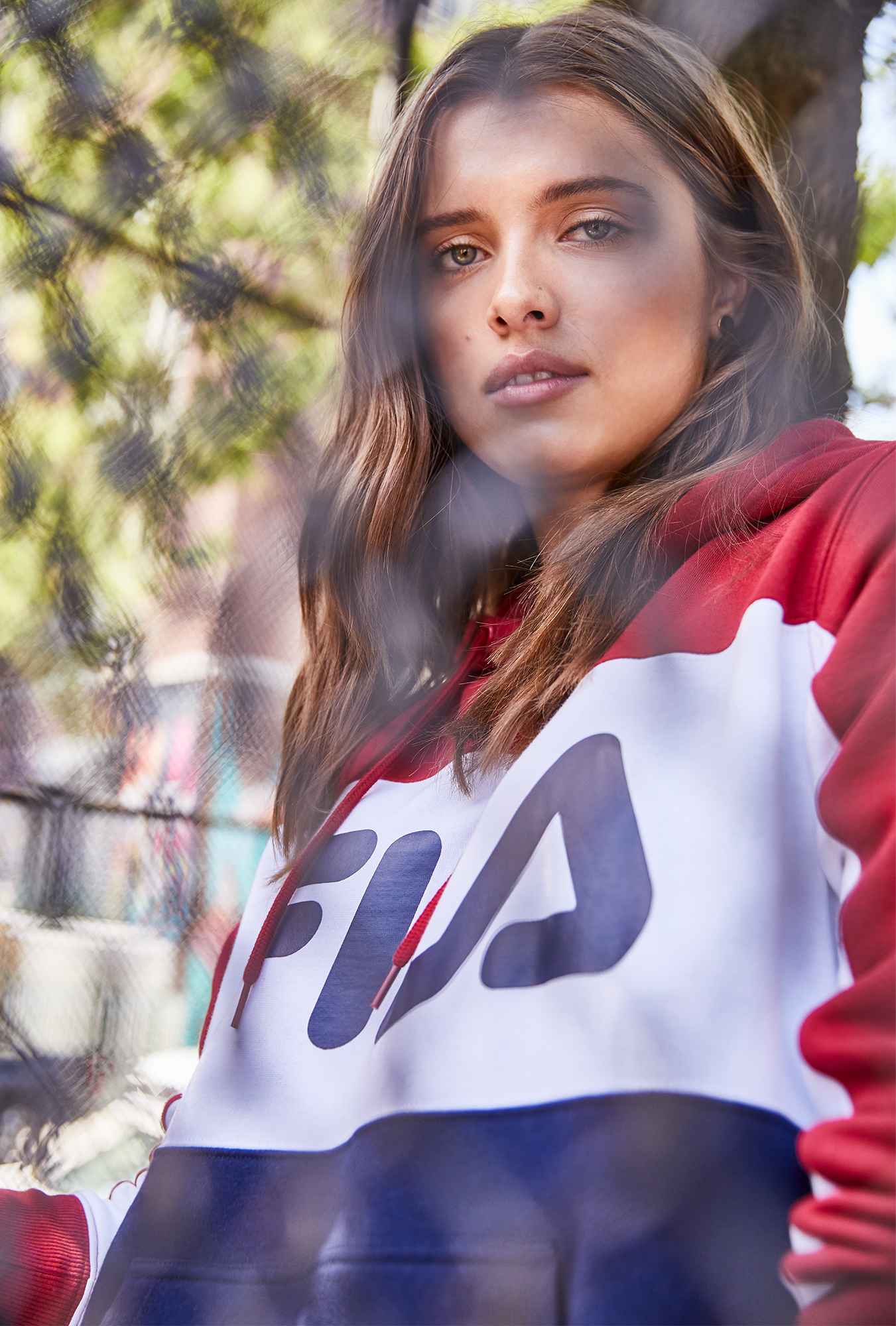 A young woman with light brown hair and blue eyes, wearing a red, white, and blue sports jacket, looking serious and outdoors near a tree.