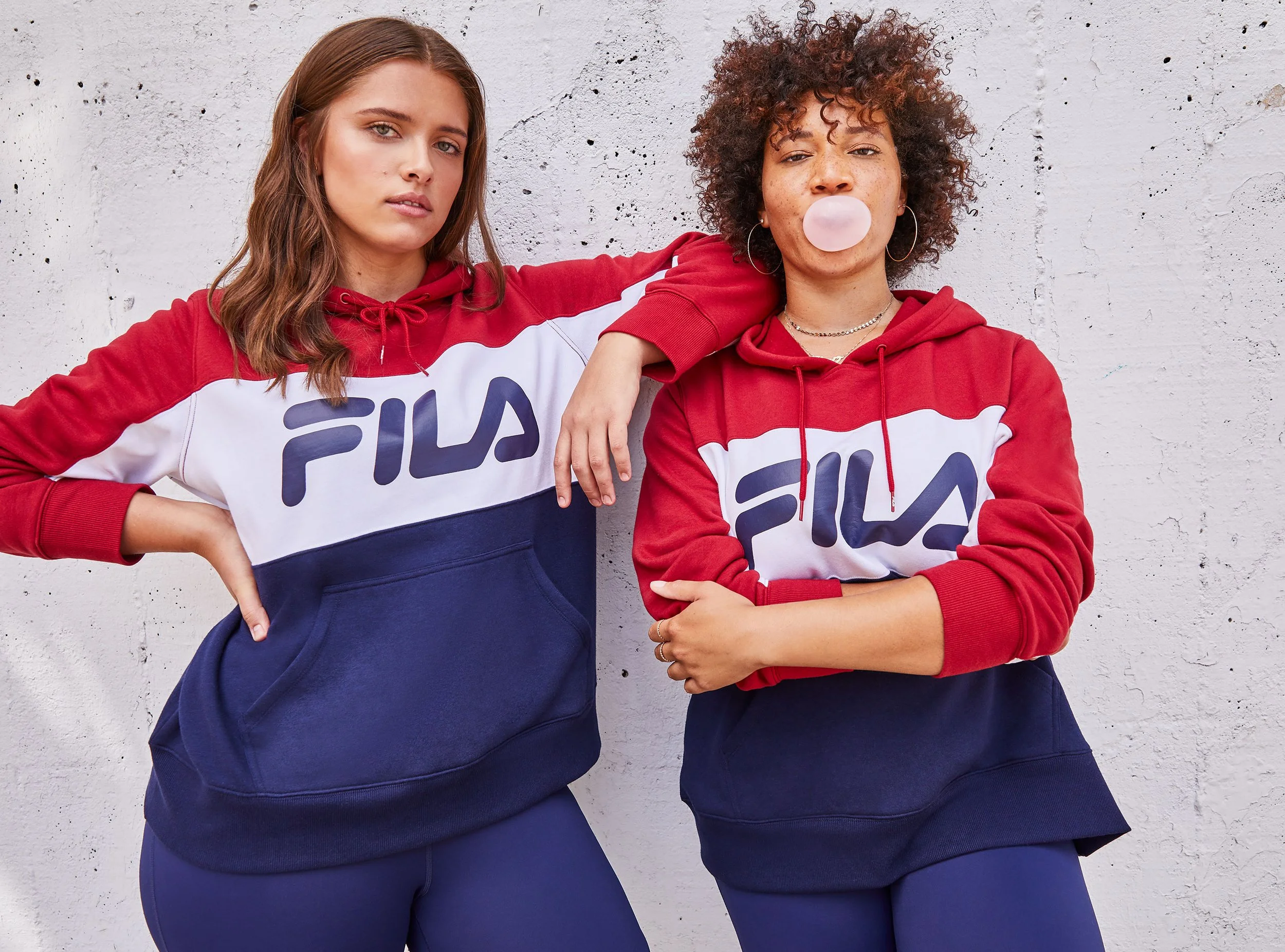 Two women standing against a white wall, wearing matching red, white, and blue Fila hoodies and navy sweatpants. One woman has long brown hair, and the other has short curly hair and is blowing a bubble with pink gum.