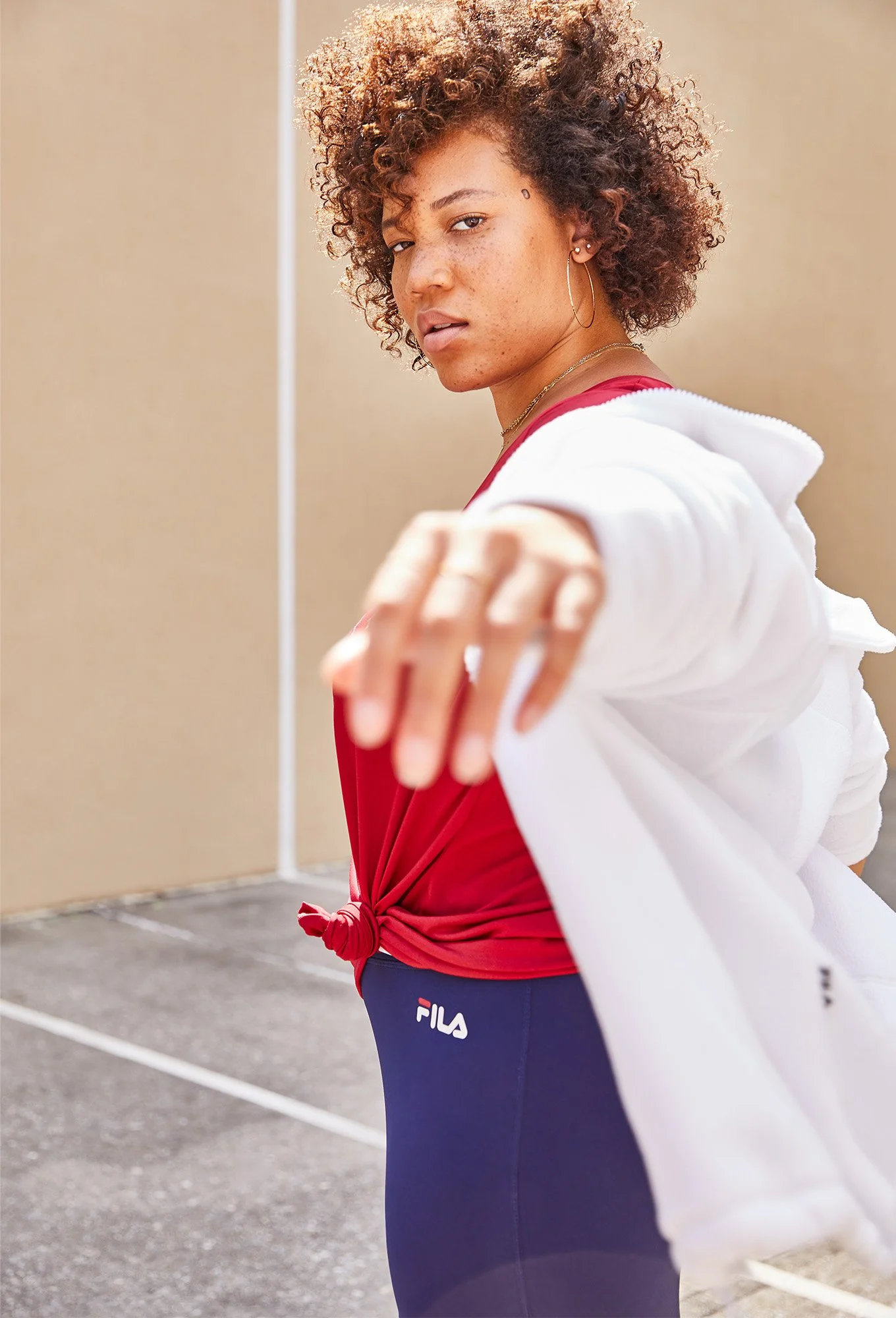 A young woman with curly hair and freckles, wearing a red shirt knotted at the waist, blue leggings with the Fila logo, and hoop earrings, standing outdoors on a tennis court, extending her hand towards the camera.