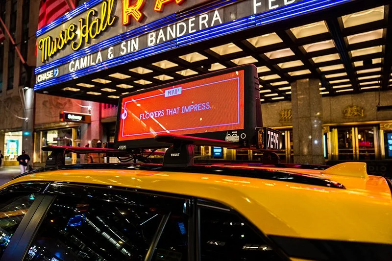 Night scene featuring a taxi cab with a rooftop digital sign displaying an advertisement that reads 'FLOWERS THAT IMPRESS.' in front of a marquee for the Music Hall of Fame, with bright lights and people walking on the sidewalk.