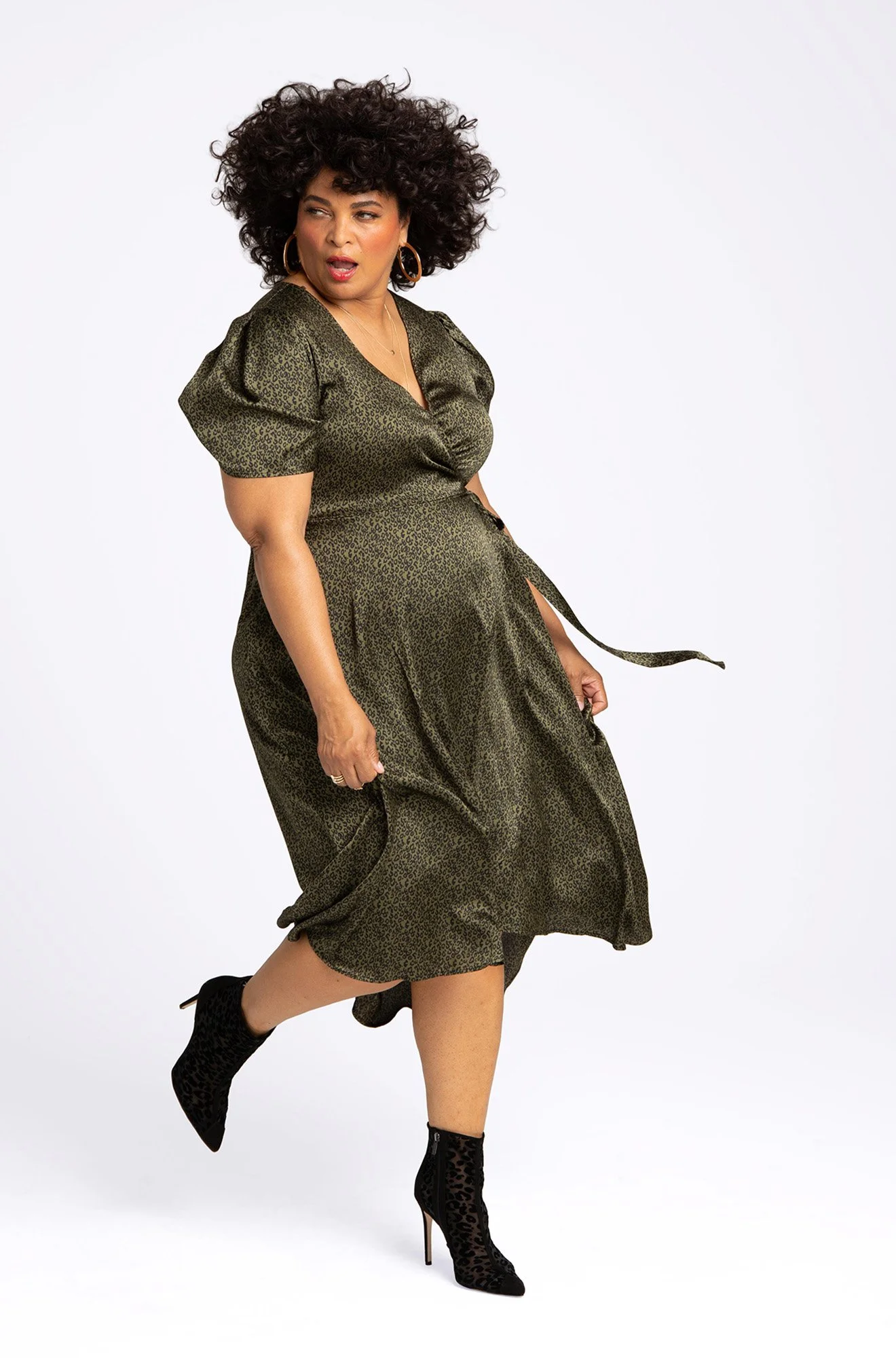 A woman with curly dark hair wearing a leopard print dress, black high-heeled leopard print ankle boots, and large hoop earrings, striking a playful pose against a plain white background.