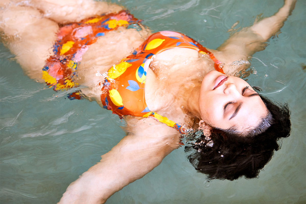 A woman floating on her back in a swimming pool with her eyes closed, wearing a colorful swimsuit with yellow, blue, and pink patterns.