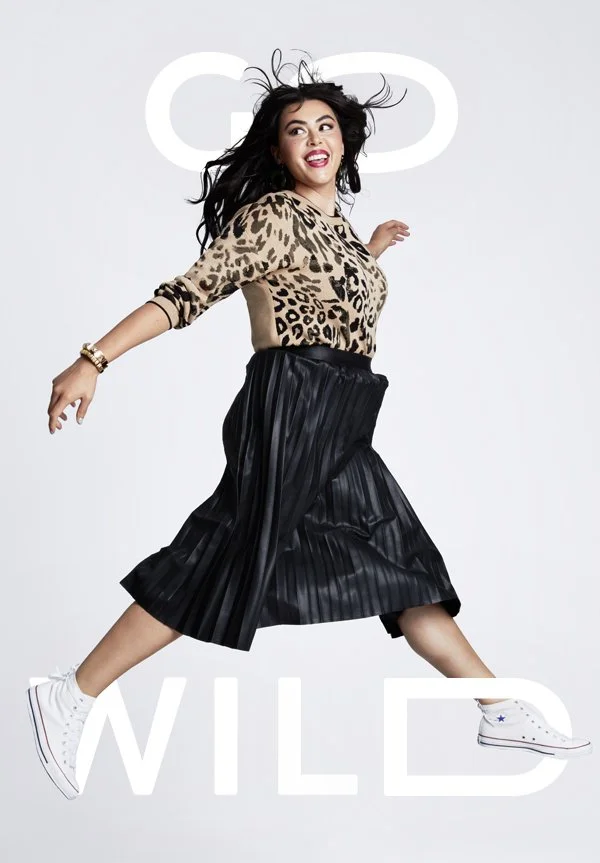 A woman in a leopard print top, black pleated skirt, and white sneakers jumping mid-air with a big smile, against a plain background with the words 'GO WILD' in the background.