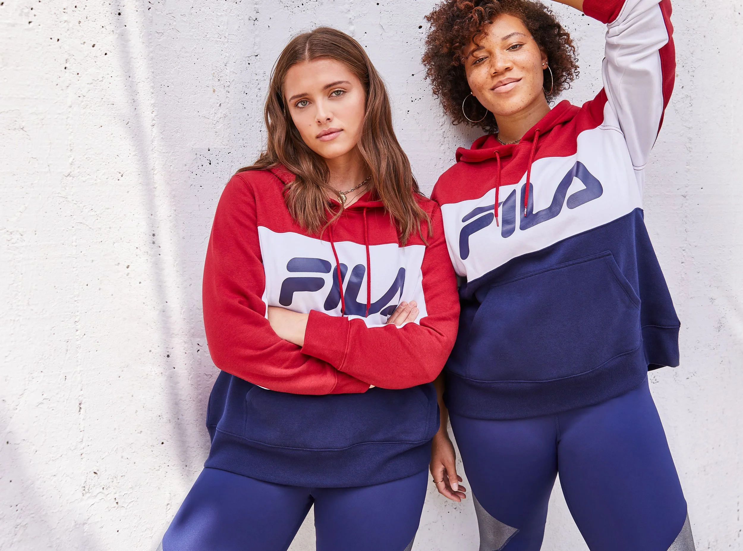 Two young women wearing red, white, and blue FILA hoodies and sweatpants standing against a white textured wall. The woman on the left has long brown hair and crossed arms, while the woman on the right has curly hair, hoop earrings, and one arm raised.