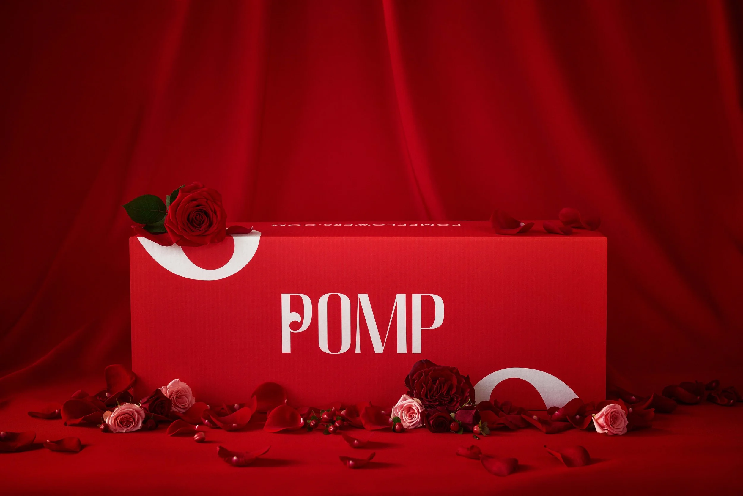 Red backdrop with a red box labeled 'PUMP' and a single red rose on top, surrounded by rose petals and flowers on the floor.