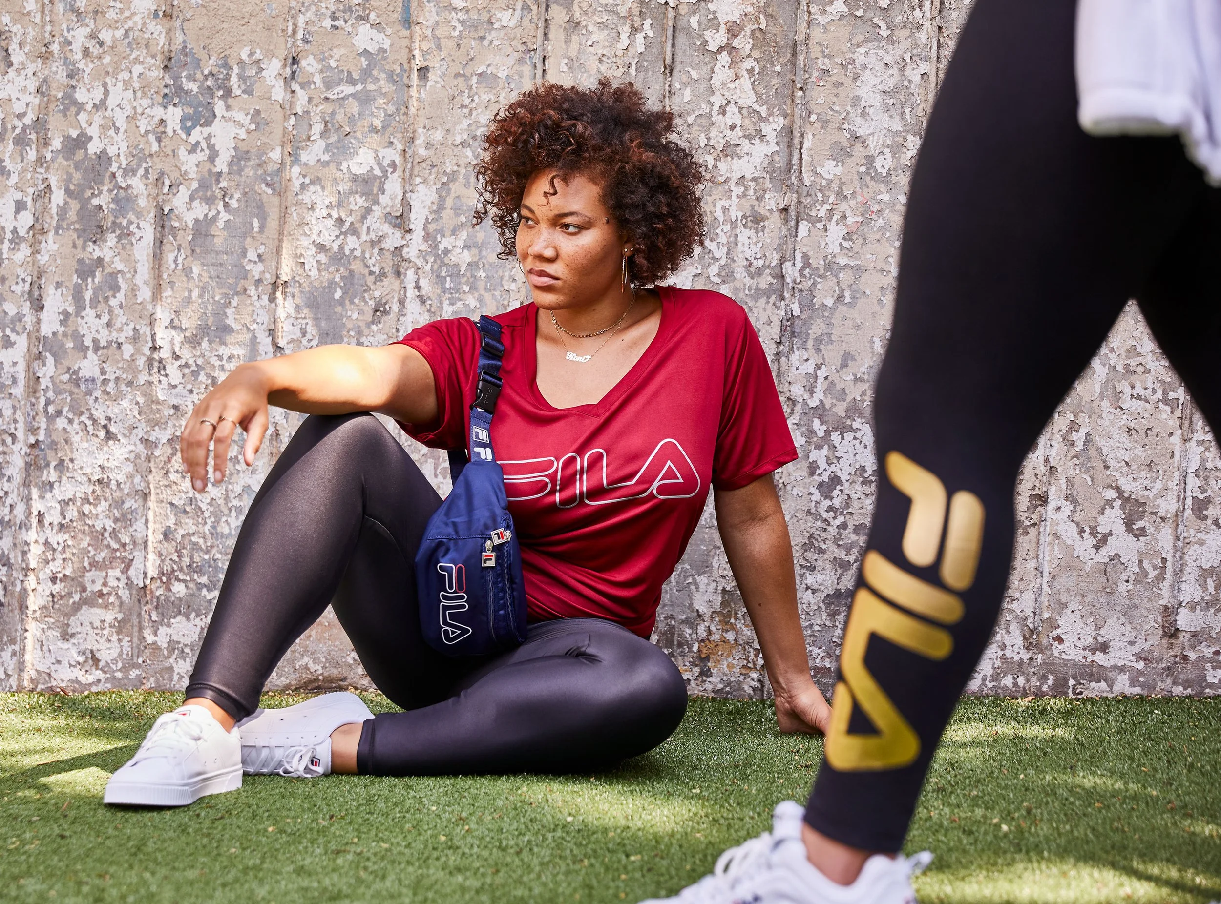 A woman with curly hair and tattoos on her arm sitting on the grass against a textured wall, wearing a red FITA shirt, black leggings, and white sneakers, while another person with black and yellow shoes stands nearby.