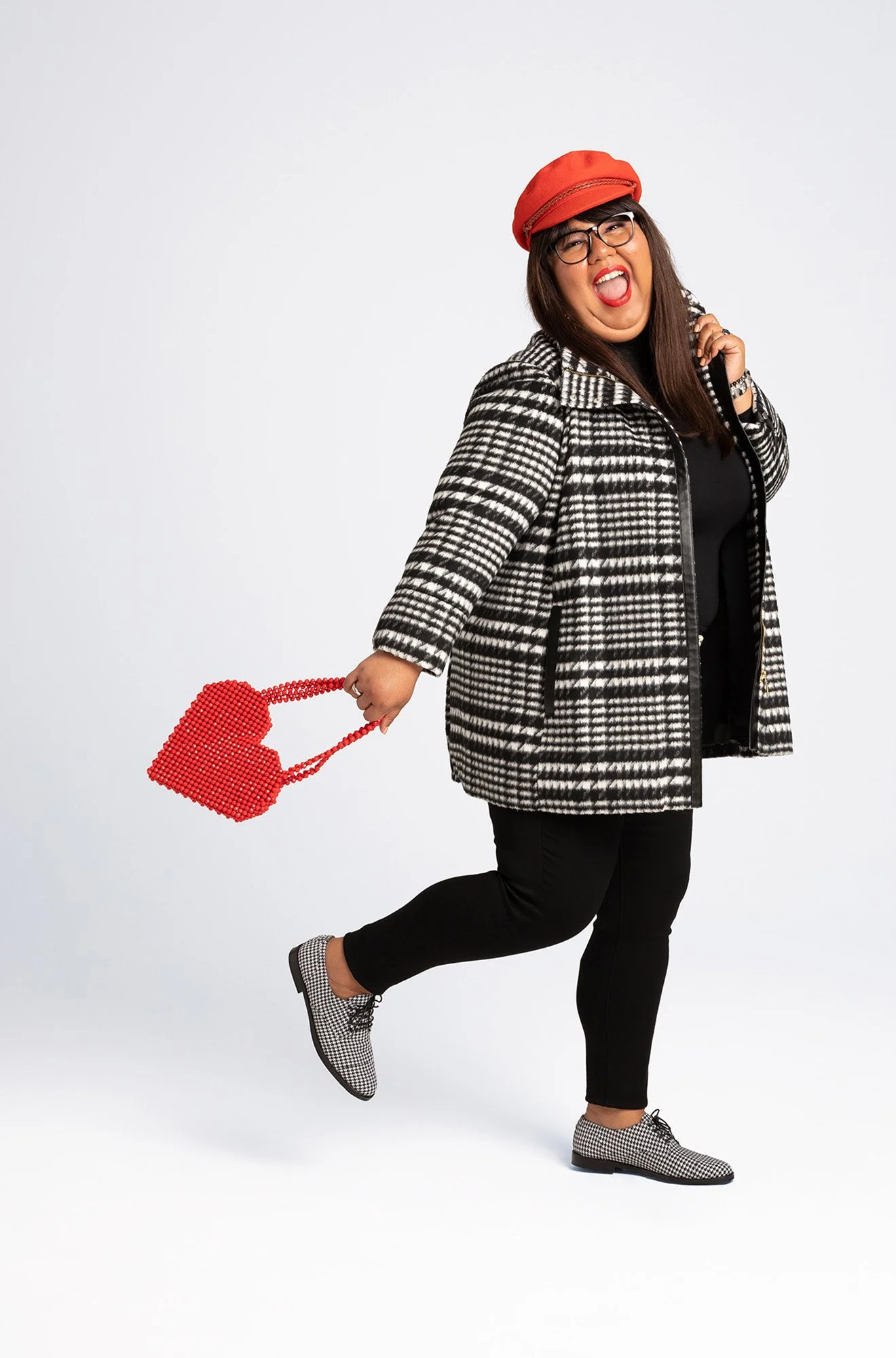 A woman wearing a checkered jacket, black pants, checkered shoes, and red hat, holding a red handbag, standing on one leg with a joyful expression.
