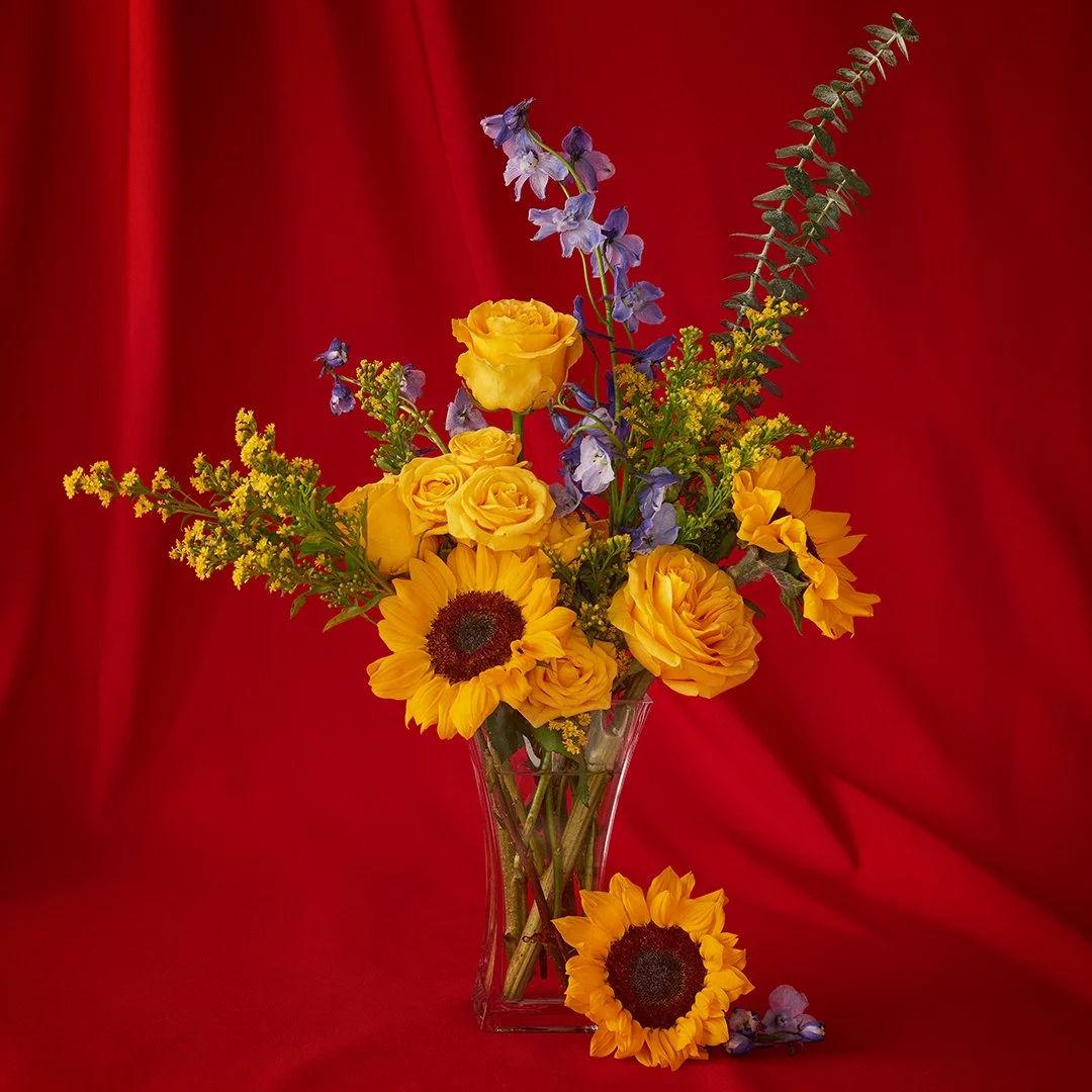 A colorful flower arrangement in a clear glass vase with yellow roses, sunflowers, blue delphiniums, and greenery set against a red fabric background.