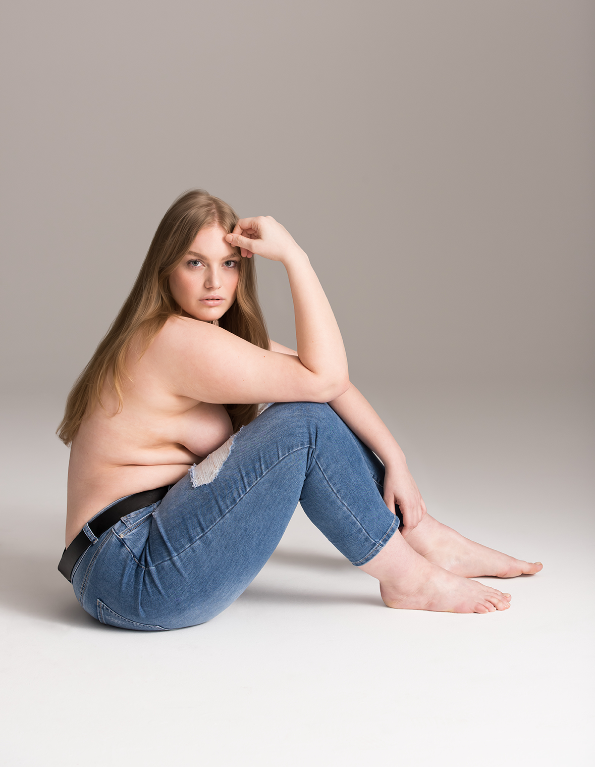 A topless young woman with long blonde hair, sitting on the floor with her legs bent, wearing ripped blue jeans, against a plain light gray background.