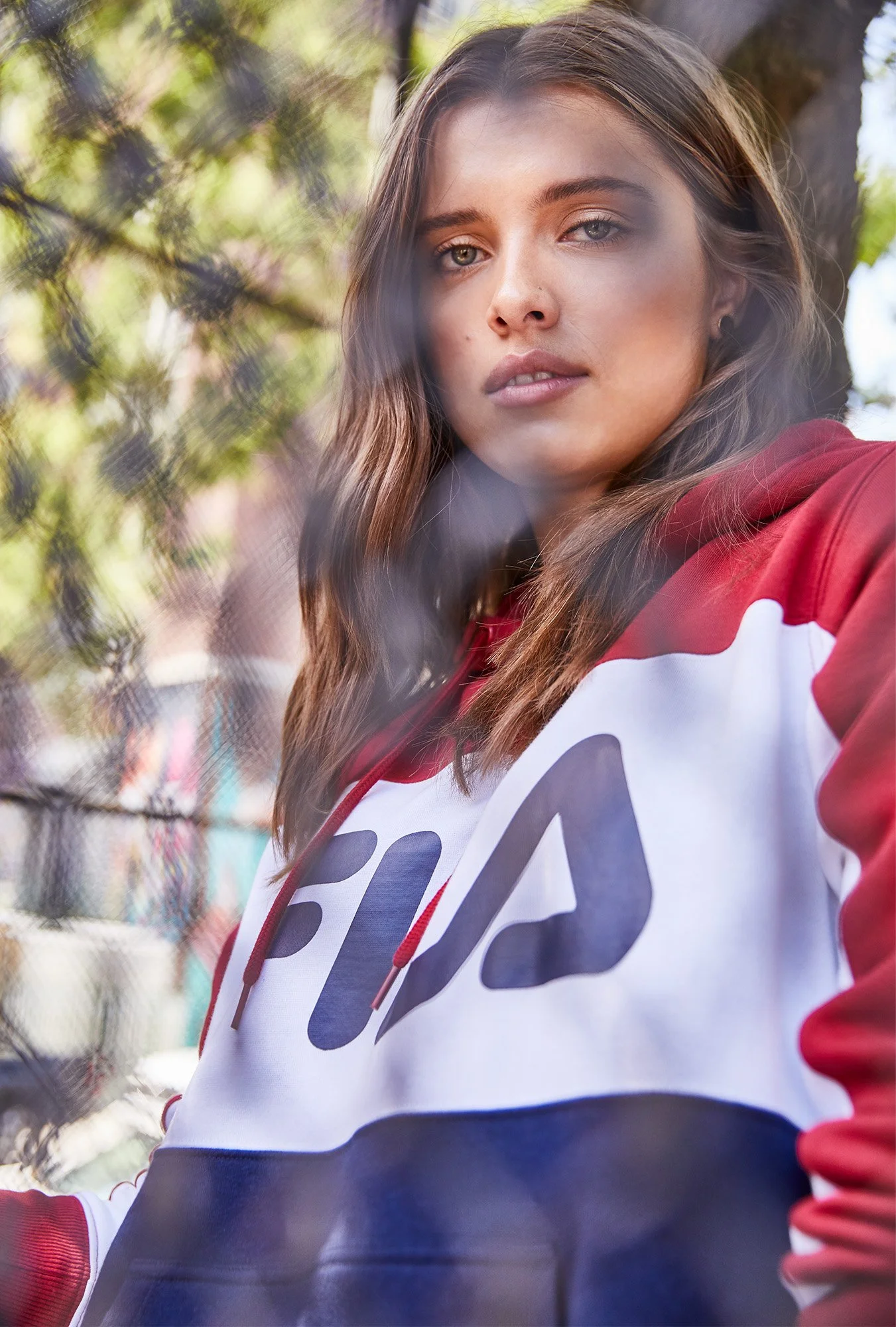 Close-up of a young woman with long brown hair and blue eyes, wearing a red and white jacket with a pacer number on her chest, outdoors, blurred background of trees and colorful fencing.