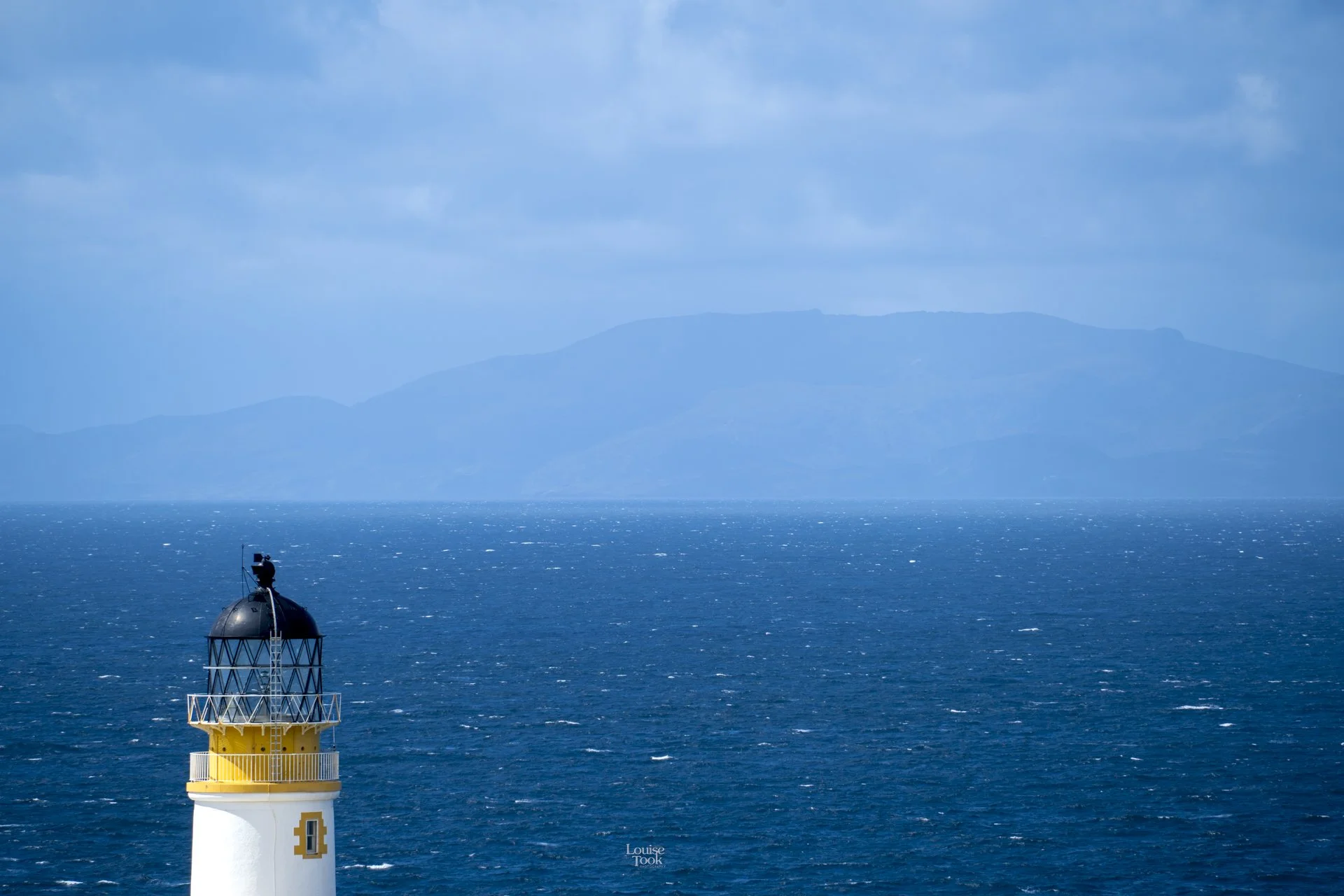 A very beautiful landscape photograph showing Neist Point lighthouse on the Isle of Skye in Scotland, looking out over the sea to the Island of Rona in the Hebrides. The image is taken on a beautiful sunny day, and the water is blue and inviting.