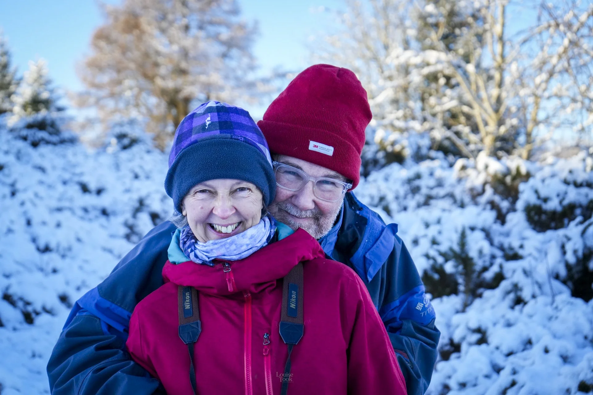 A couple hug and smile at the camera, they are dressed in warm winter jackets and hats as the scenery is snowy. They look very happy.