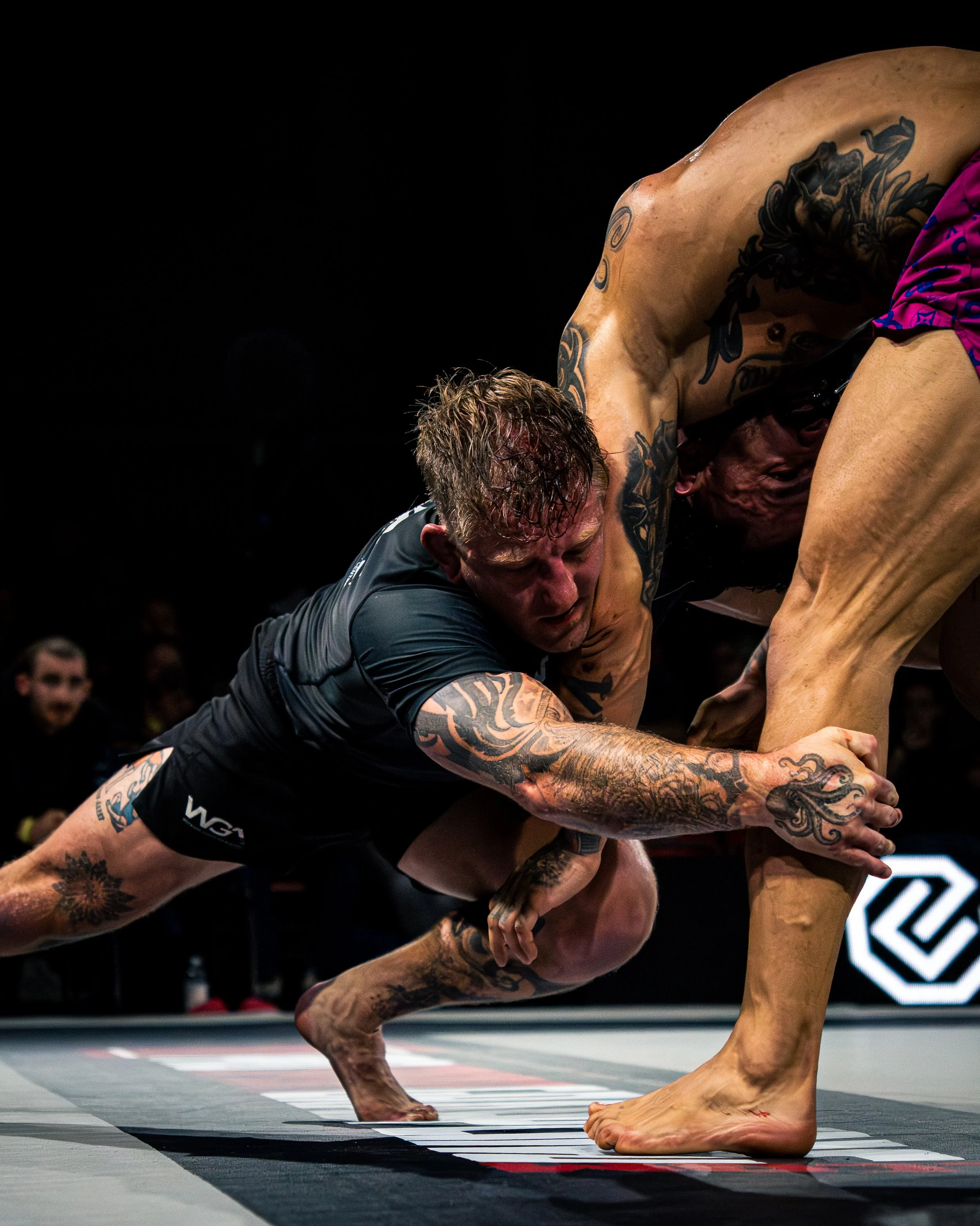 Mixed martial arts fighters grappling during a match, with one fighter in black shorts and the other partially visible, on a mat with match markings, in an arena with spectators.