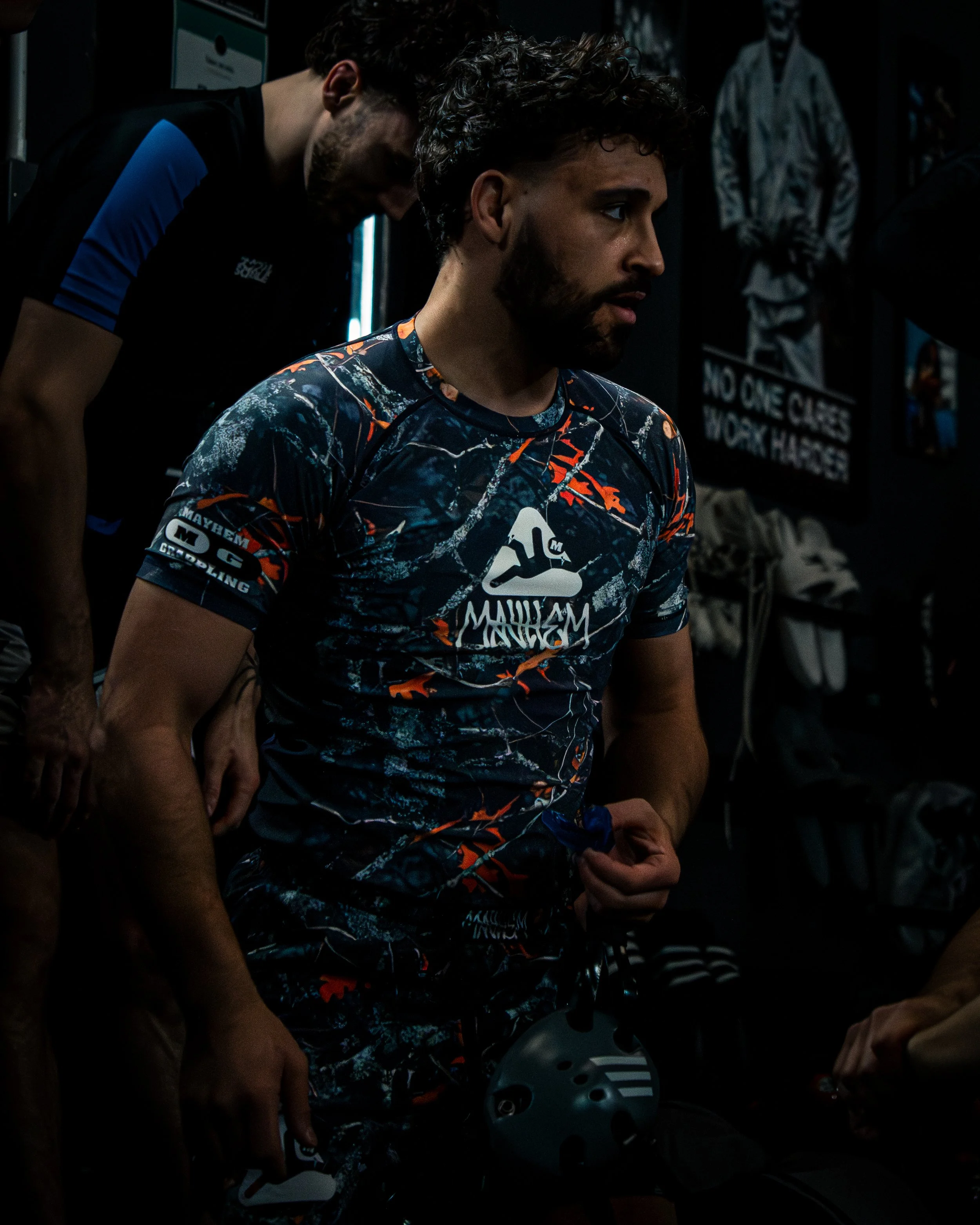A young man with dark curly hair and a beard in a motocross jersey holding a motorcycle helmet, in a dimly lit room with posters on the wall.