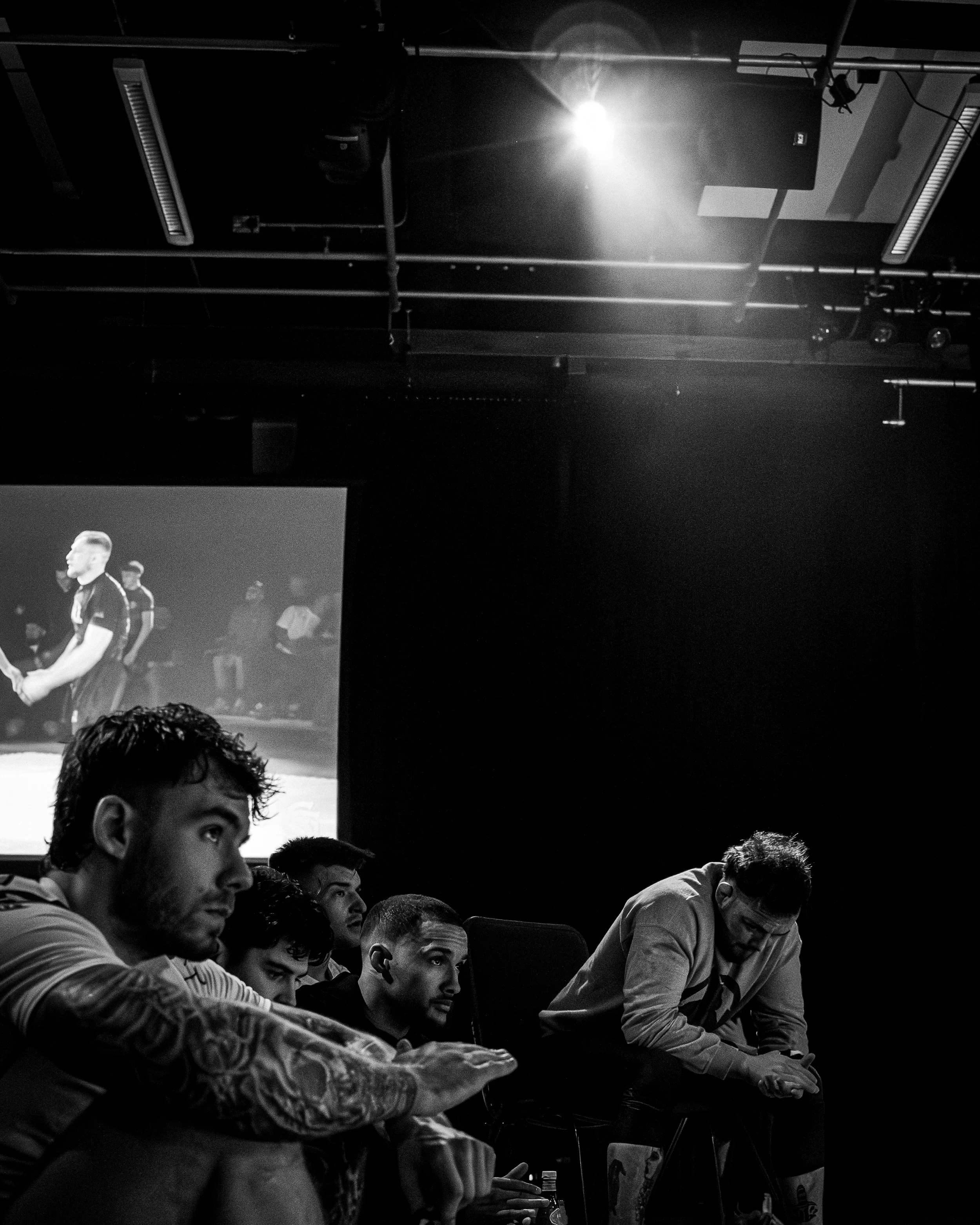 Black and white photo of a group of people sitting at tables, with some looking down and some looking forward. The background features stage lighting, a projector screen with a person speaking, and ceiling fixtures.
