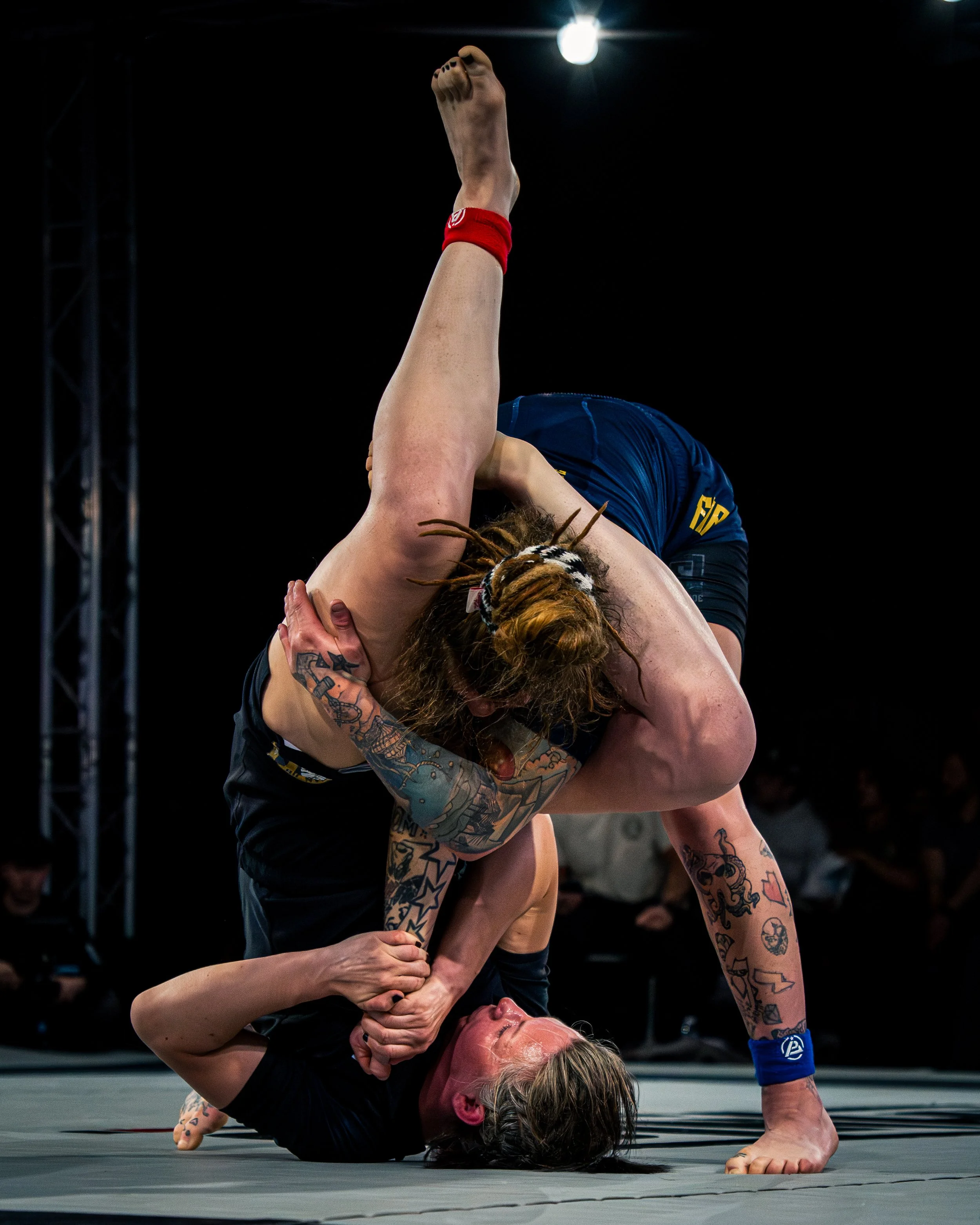 Two female wrestlers are engaged in a match, with one on top of the other on a wrestling mat. The wrestler on top has her arm raised and is wearing a red wristband, and the one on the bottom is holding onto her opponent. Both women have tattoos and a