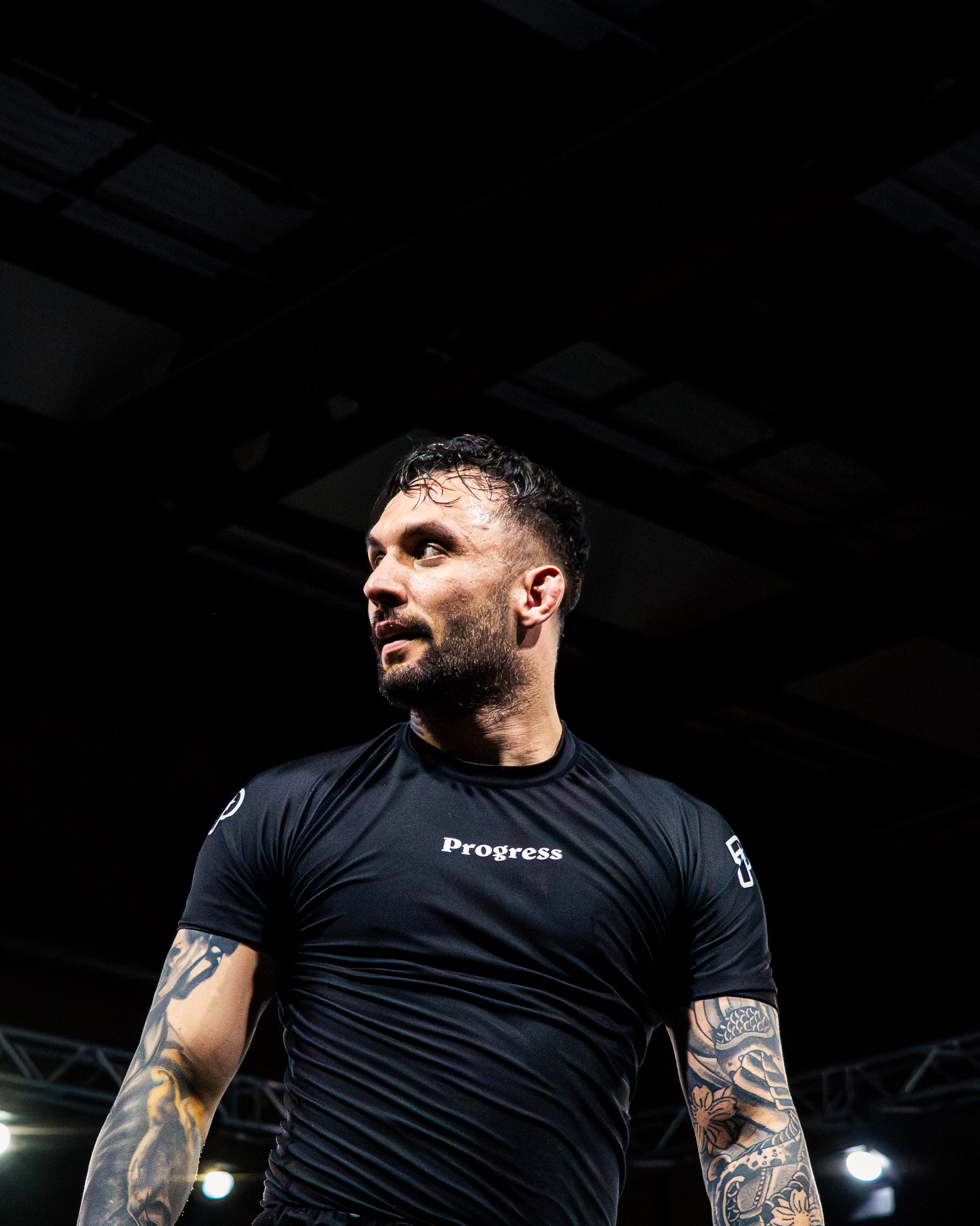 A man with tattoos on his arms wearing a black t-shirt with the word "Progress" printed on it, standing under a dark ceiling with visible beams, looking to his left.