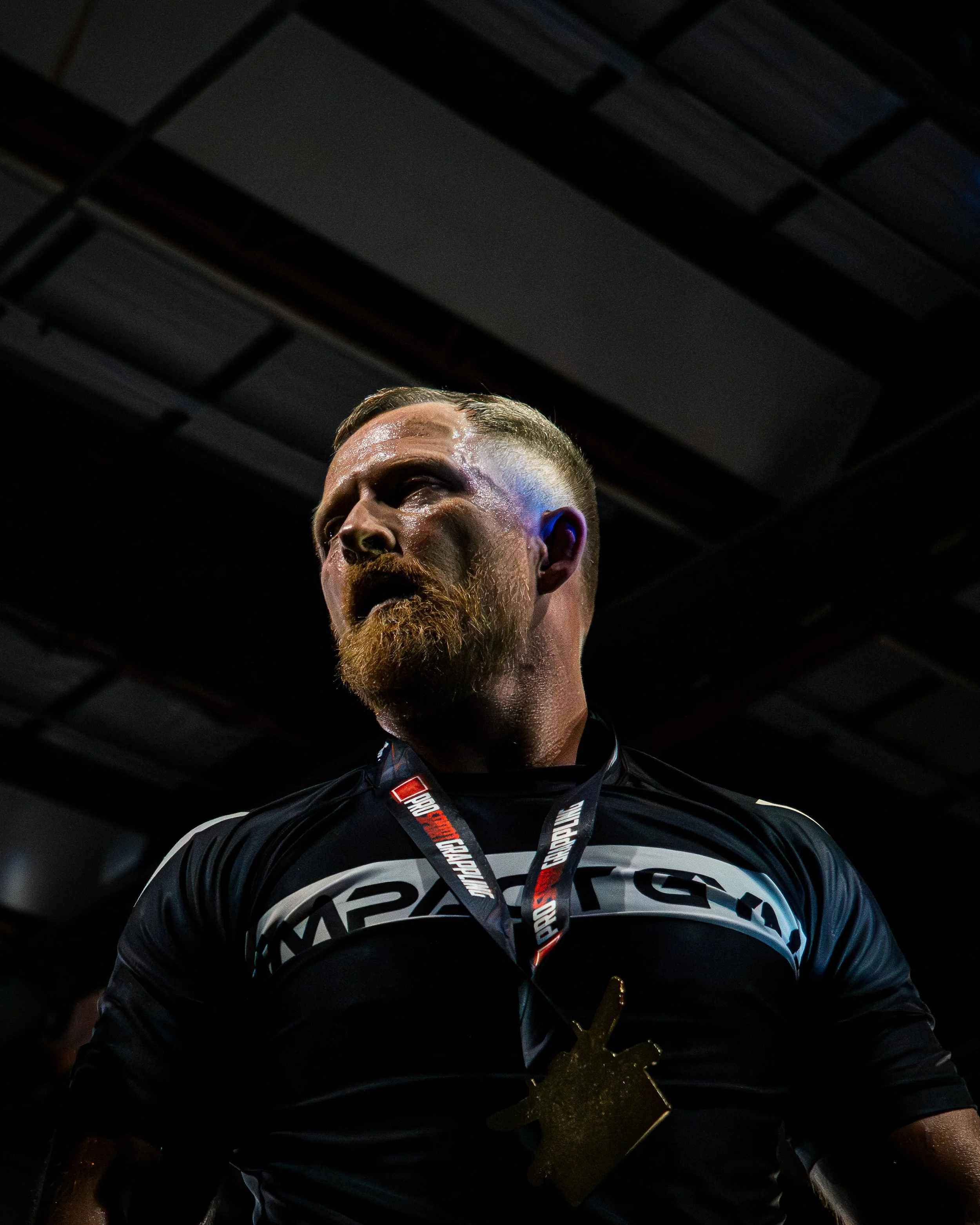 A man with a beard, wearing a black and white sports jersey and a winner's medal around his neck, looking slightly to the right, with a dark ceiling in the background.